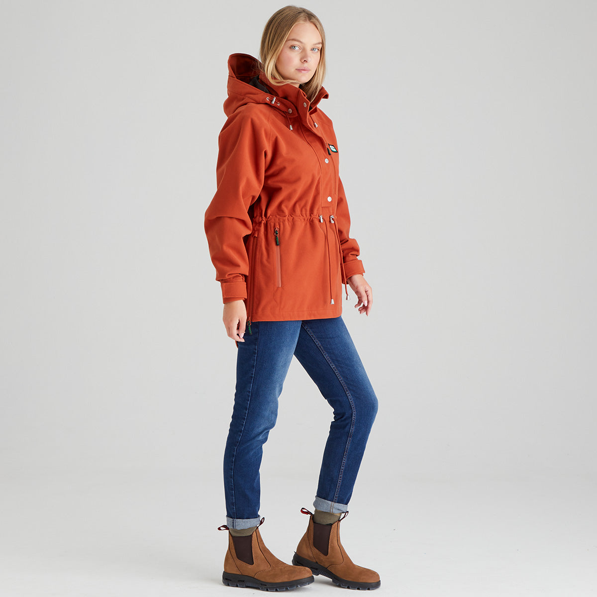 Person wearing an orange jacket, blue jeans, and brown boots on a light grey background