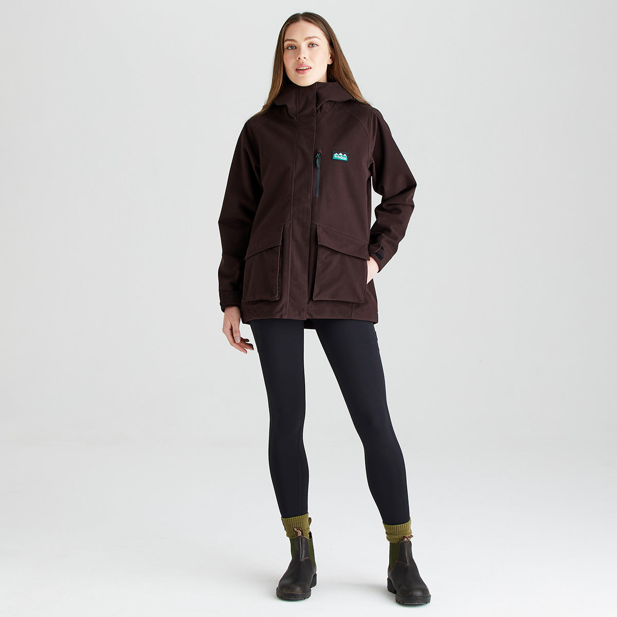 Dark brown hooded outdoor jacket with high collar, front zip and chest pocket, full body front view worn with black leggings and ankle boots in a grey studio background.