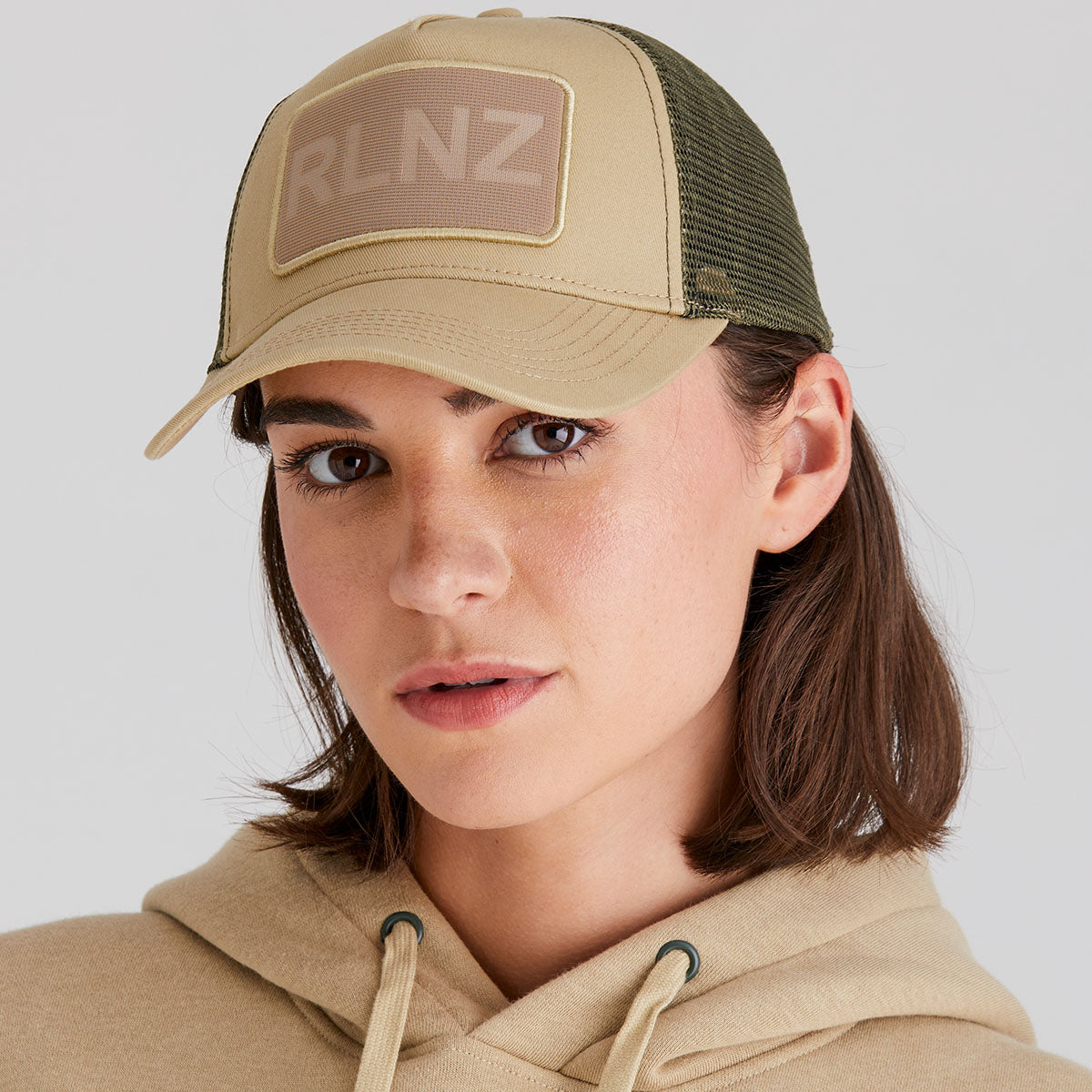 Person wearing a beige cap with a patch and matching hoodie on a light gray background