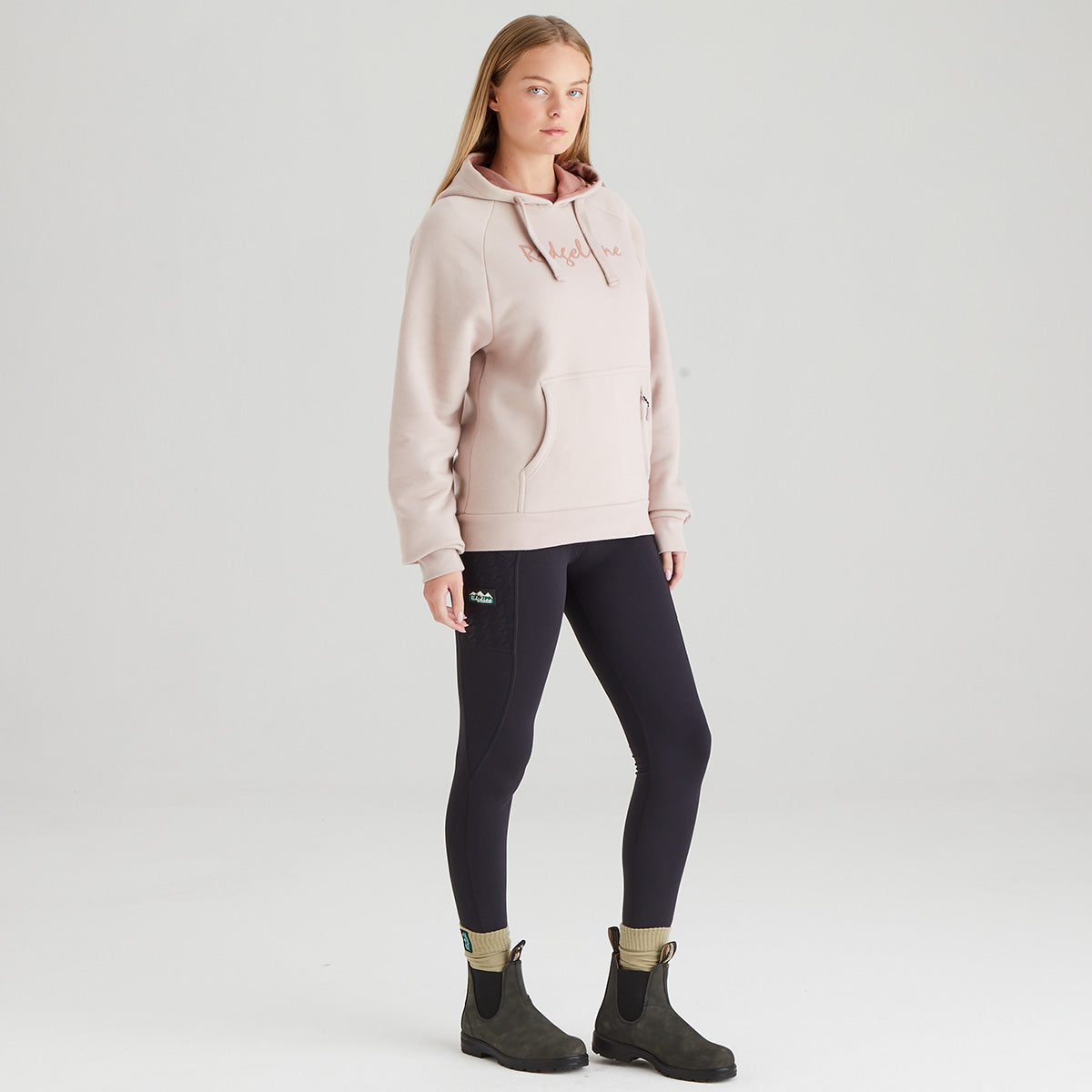 Woman wearing a light pink hoodie, black leggings, and dark boots on a white background