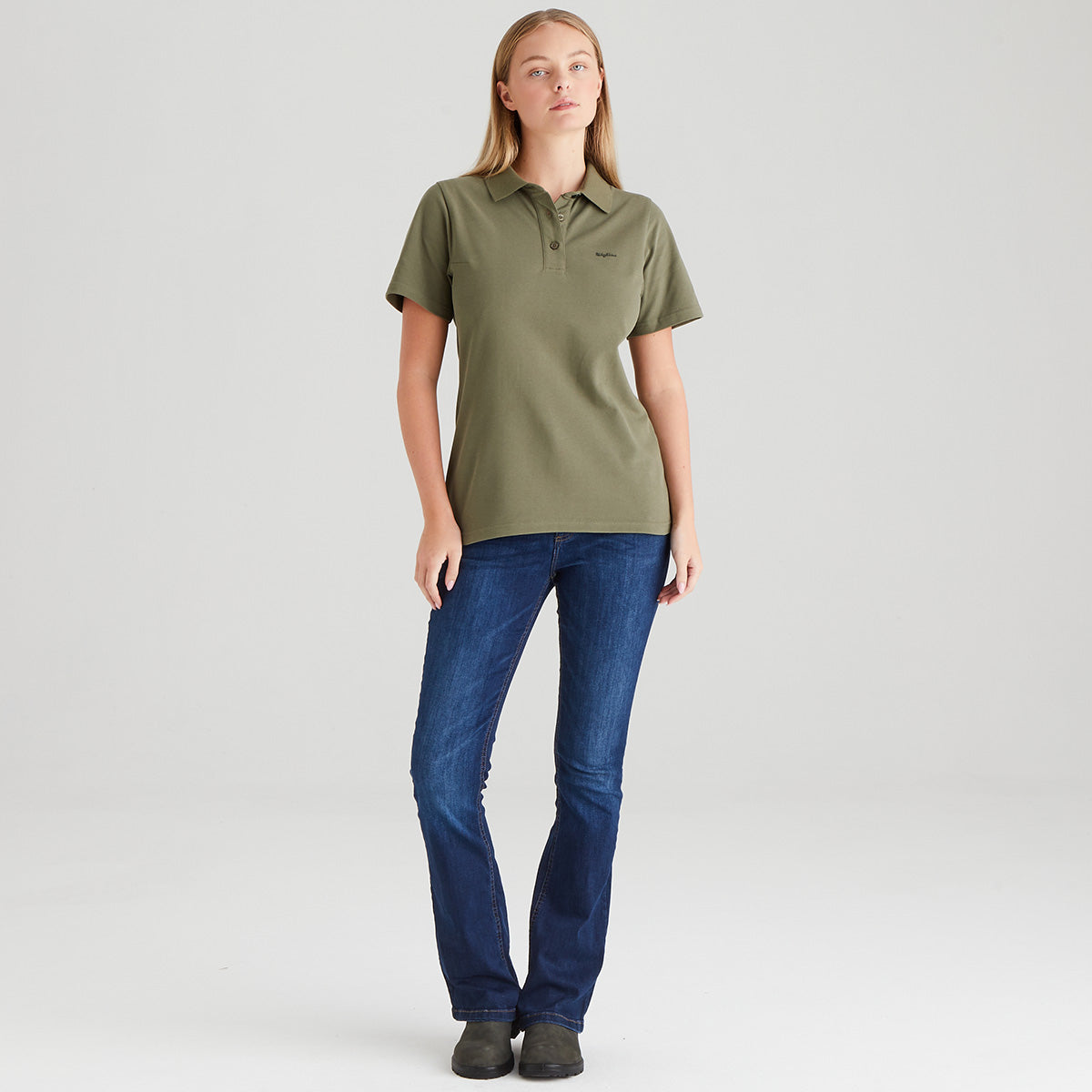 Woman wearing an olive green polo shirt and blue jeans on a light gray background