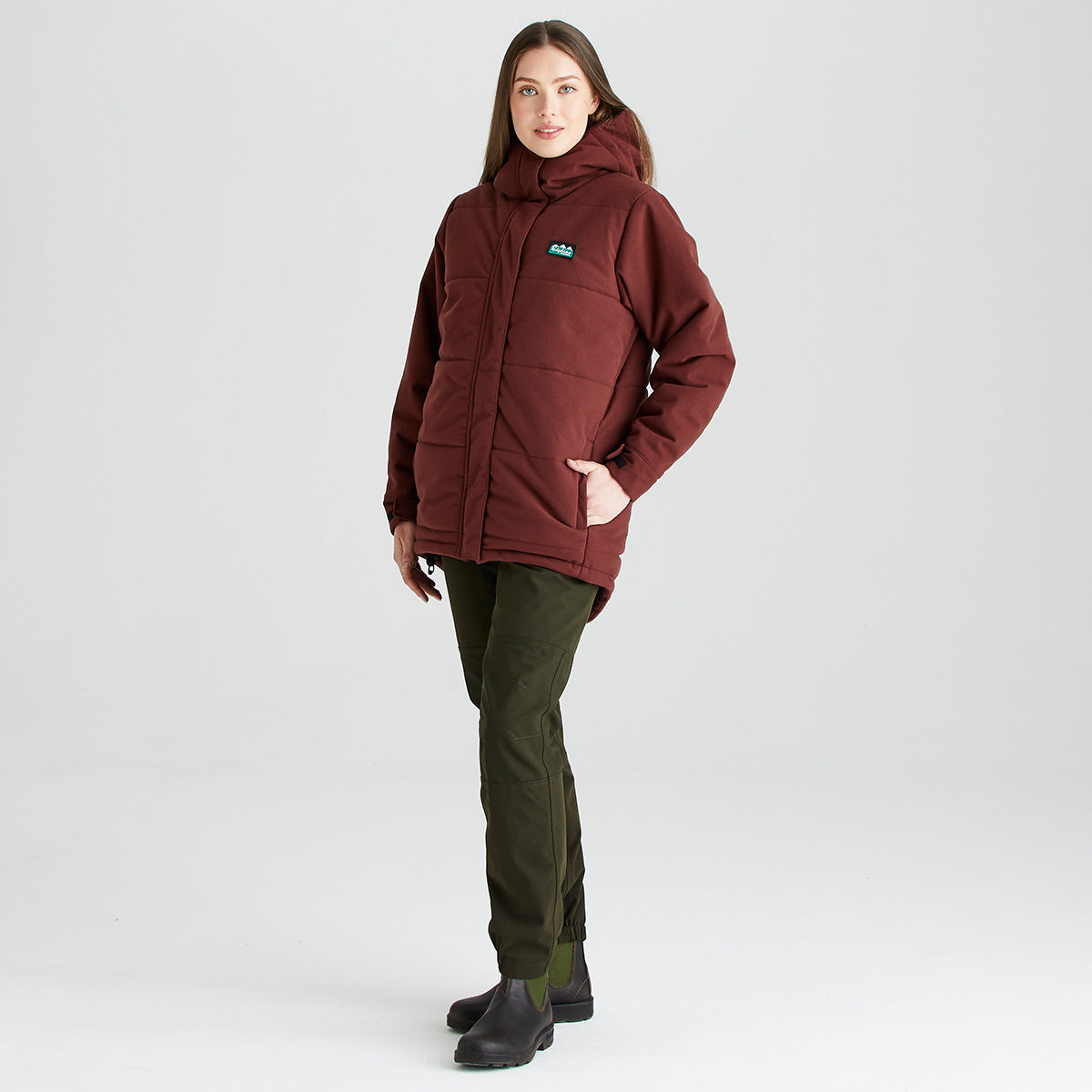 Person wearing a Ridgeline insulated jacket in burgundy with a hood, full-length concealed zip, padded construction and side pockets, paired with olive trousers and outdoor boots, shown against a light grey studio background.