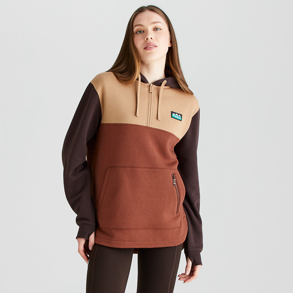 Front view of a Ridgeline colour-block hoodie in beige, rust brown, and dark brown tones, worn with dark brown leggings. The hoodie features an adjustable drawstring hood, a partial button placket at the neckline, and a large kangaroo-style front pocket with a vertical zipped pocket on one side for secure storage. A Ridgeline logo patch is displayed on the left chest. Photographed against a plain light-grey studio background.