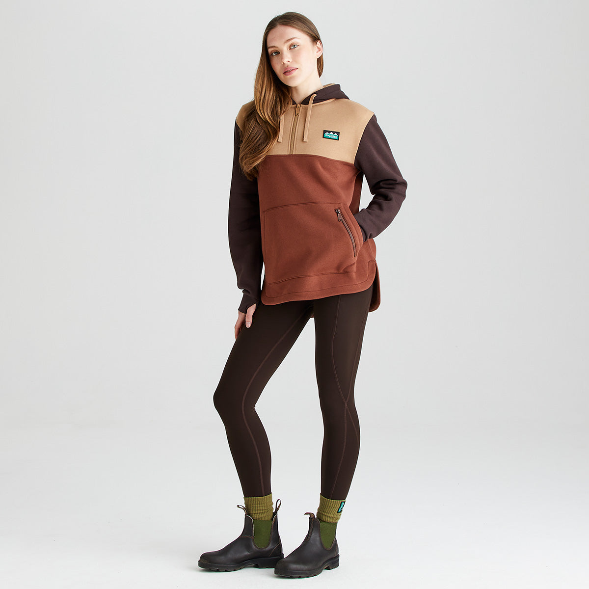 Full-length studio image of a person wearing a Ridgeline colour-block hoodie in beige, rust brown, and dark brown tones, paired with dark brown leggings, olive-green socks, and black ankle boots. The hoodie features an adjustable drawstring hood, a partial button placket at the neckline, a large kangaroo-style front pocket, and a vertical zipped pocket for secure storage. A Ridgeline logo patch is displayed on the left chest. Photographed against a plain light-grey background.
