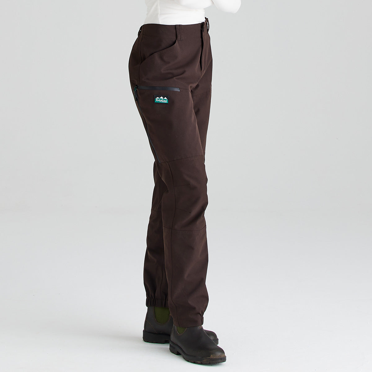 Side view of dark brown waterproof trousers shown against a light gray studio background. The trousers feature a button-fastening waistband, belt loops, articulated leg panels, a waterproof zip pocket, and a small logo patch on the upper leg. Elasticated ankle cuffs provide a secure fit over black ankle boots. The image highlights the side profile, durable construction, and practical outdoor performance features of the trousers.