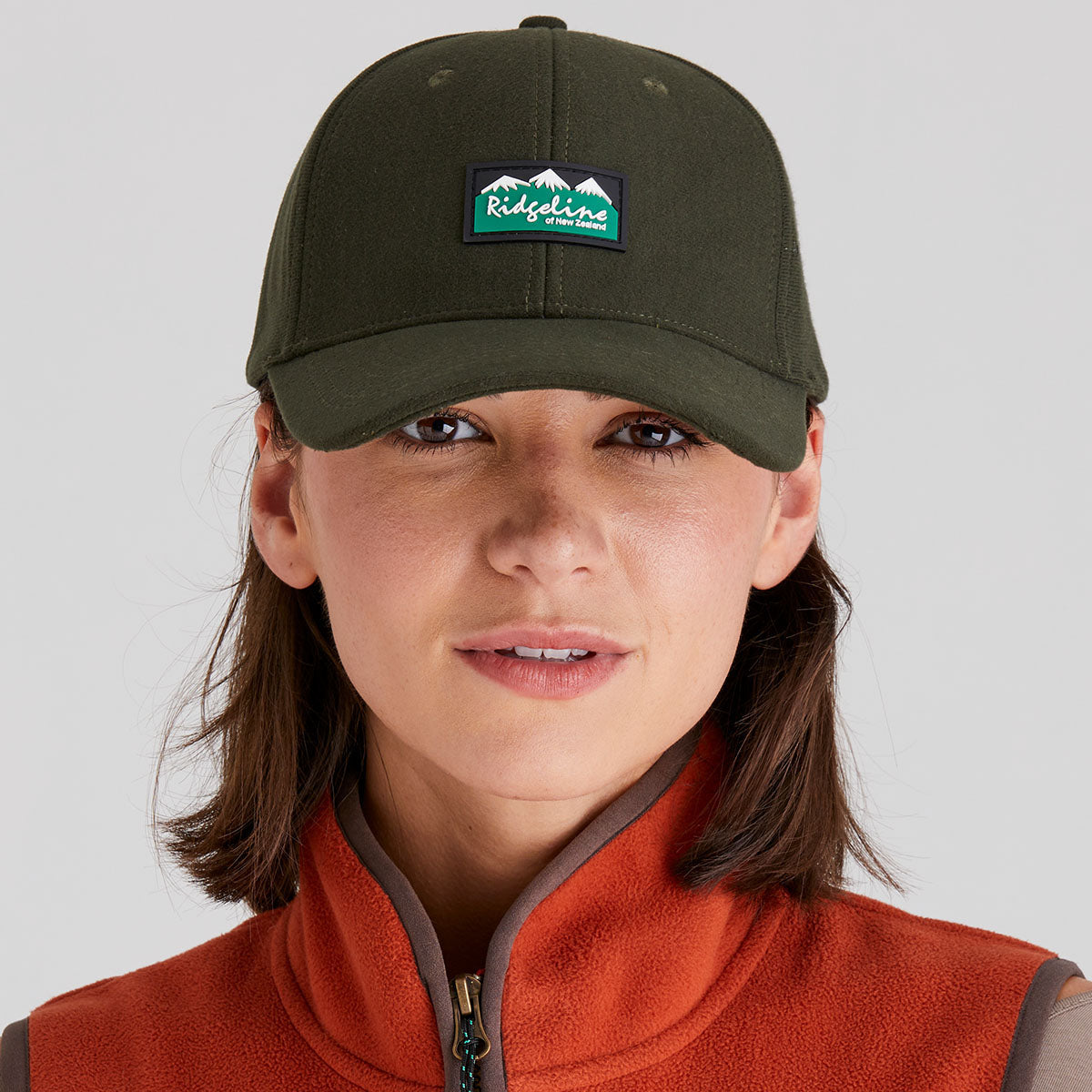 Person wearing a green cap with a logo and an orange jacket on a gray background