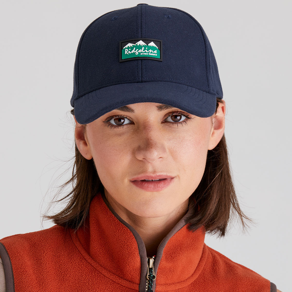 Person wearing a navy cap with a logo and an orange jacket on a gray background