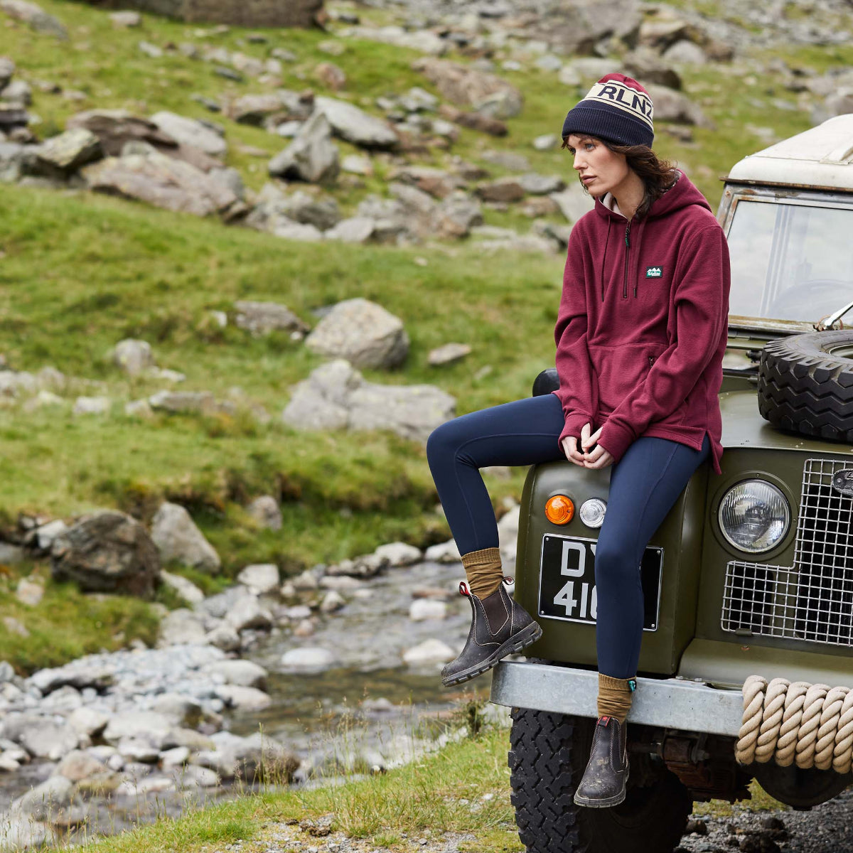 Woman wearing a red coloured fleece hoodie with a hood, front pouch pocket, and thumb loops, displayed against a plain background. Ridgeline Clothing Logo on Chest. Sitting on a car with rocks in the backround