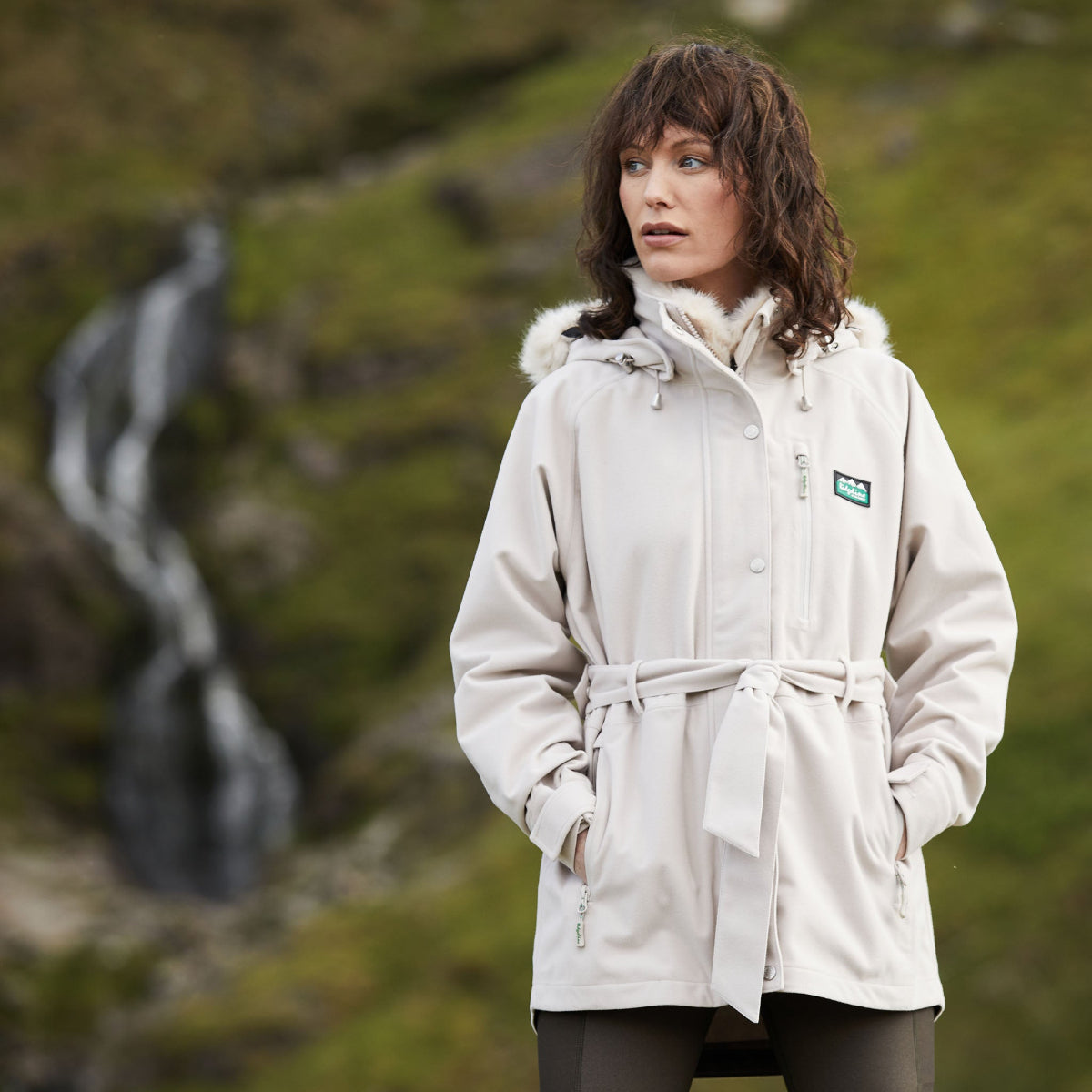 Woman wearing a beige Monsoon Nordic jacket with a faux-fur collar, belt at the waist, front zip, and a deep hood. With blurred mountain background