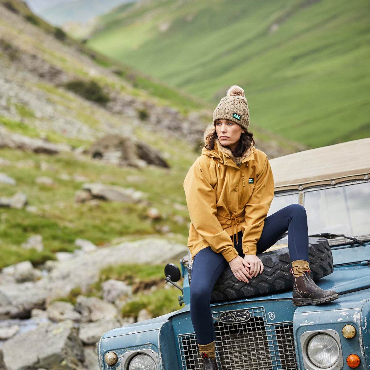 woman wearing a yellow Monsoon Nordic Smock jacket sitting on the hood of a jeep. Mountains in the background