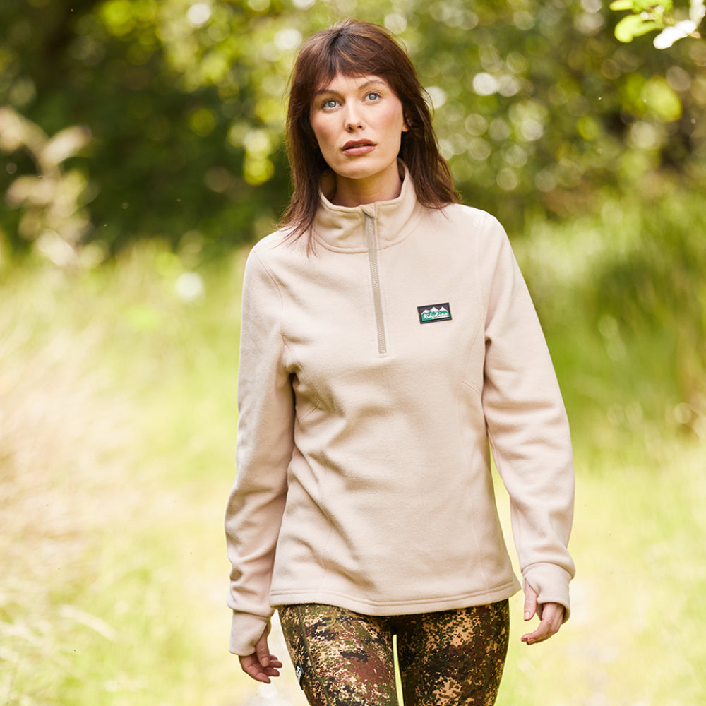 Women wearing a Cream fleece sweatshirt with a half zipper and a Ridgeline Clothing logo on the chest. Standing in green area with trees behind