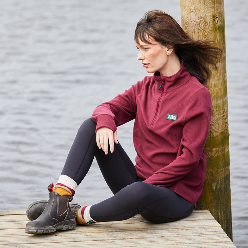 Woman wearing a red fleece sweatshirt with a half zipper and a Ridgeline Clothing logo on the chest. Sitting on decking next to a river