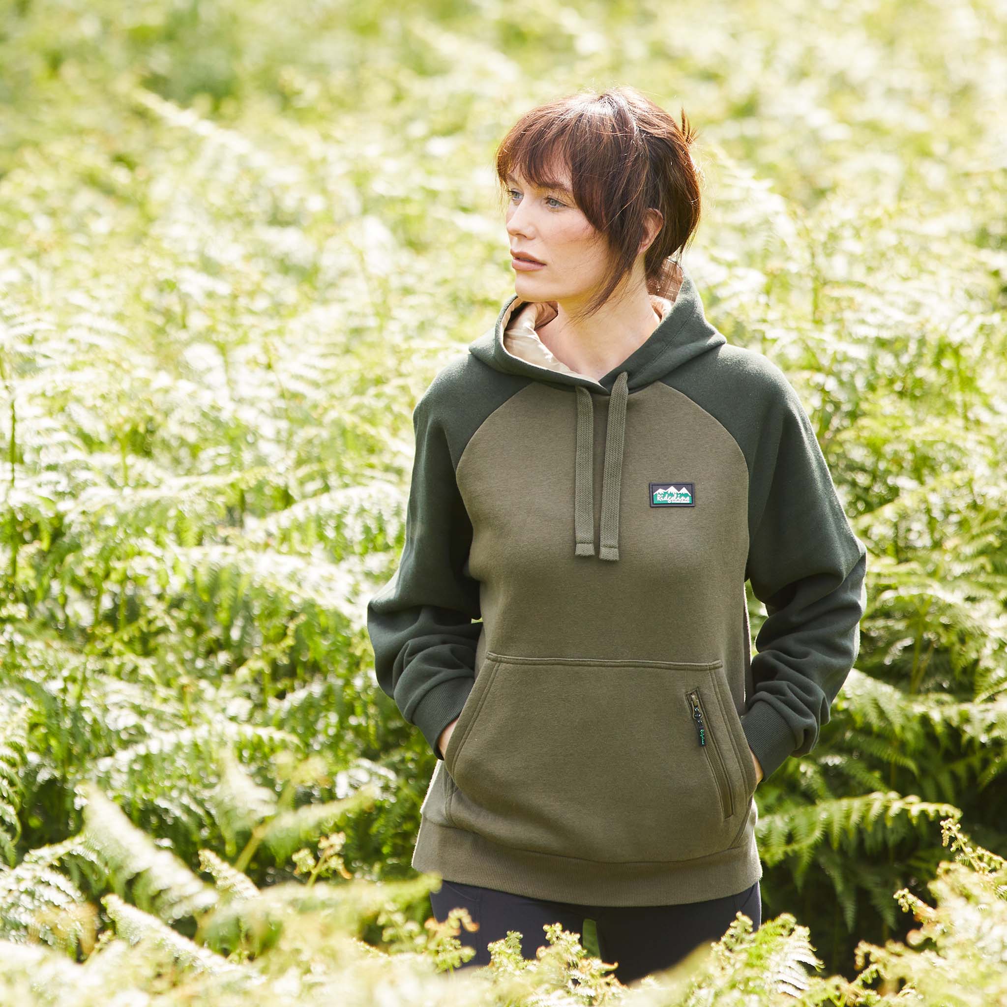 Woman wearing a green and brown hoodie with a kangaroo pocket and a small zipped valuables pocket, featuring a Ridgeline Clothing logo on the chest. Walking through a field of plants