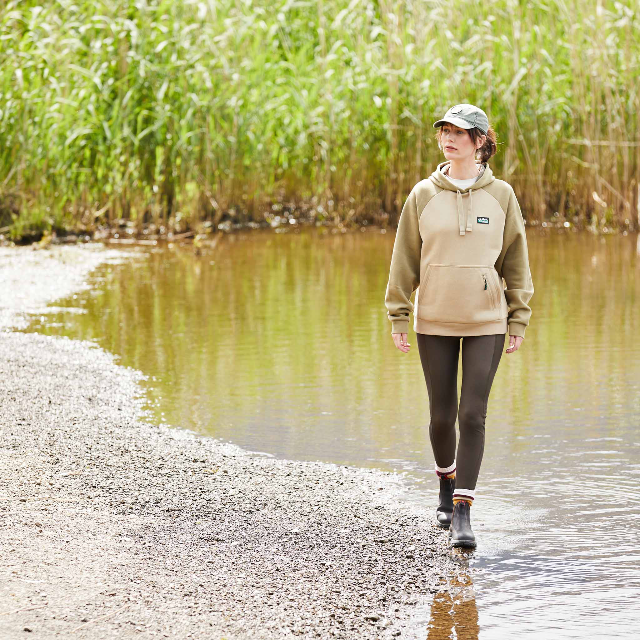 Woman wearing a beige and brown hoodie with a kangaroo pocket and a small zipped valuables pocket, featuring a Ridgeline Clothing logo on the chest. Walking alongside a river with high grass in the background