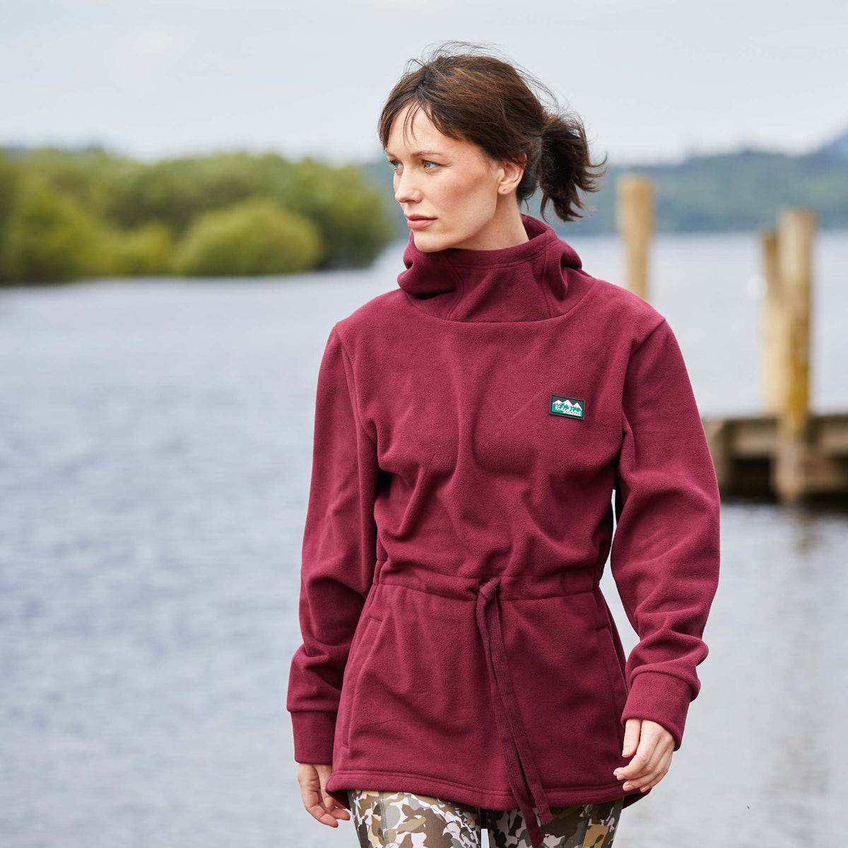 Woman wearing a red fleece hoodie with a high collar, hood, and two pockets, displayed in an outdoor setting with a river and trees in the background