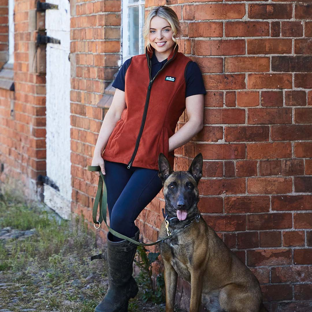 Woman in orange vest and blue pants standing next to a dog against a brick wall.