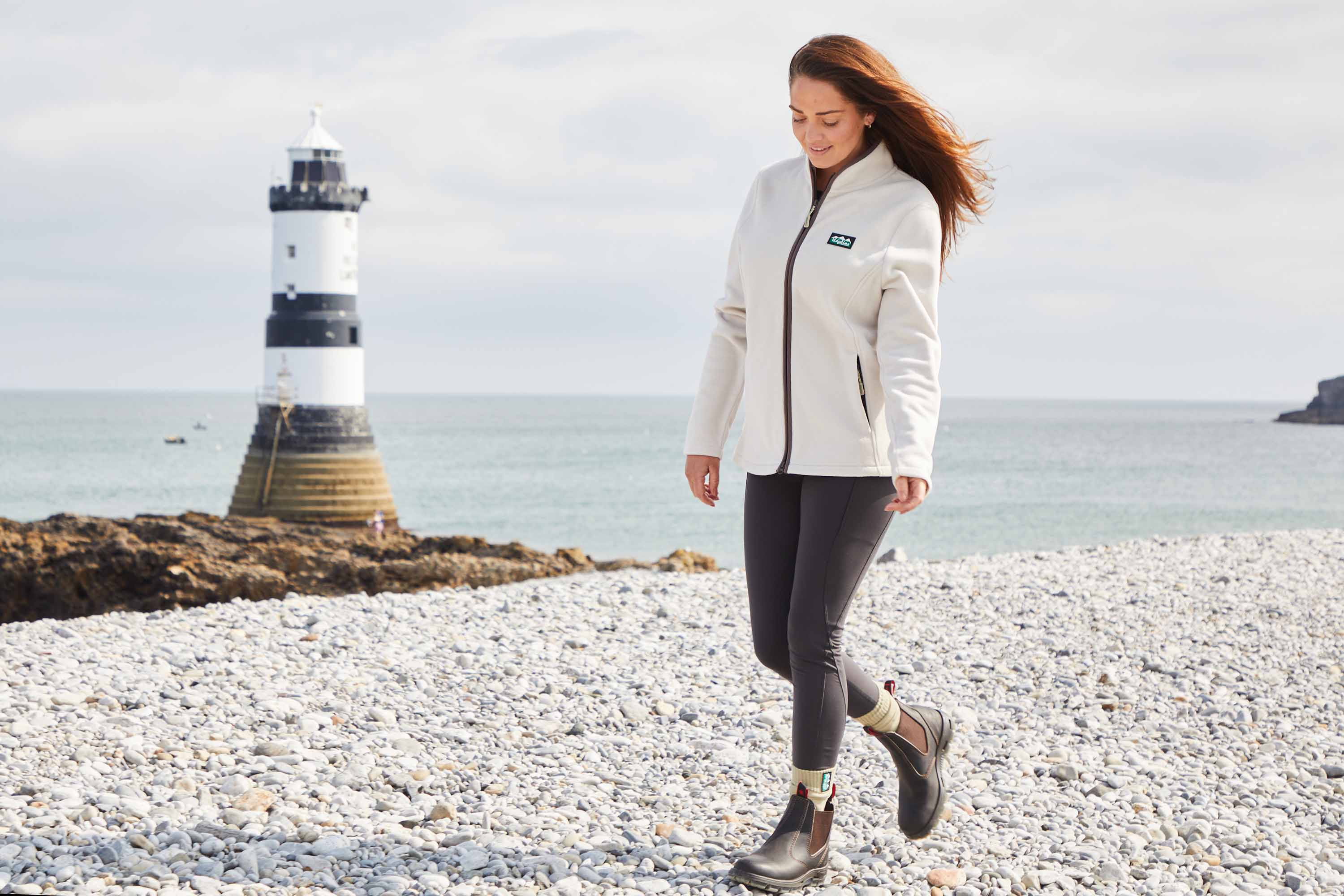 Woman walking on a pebbly beach with a lighthouse in the background