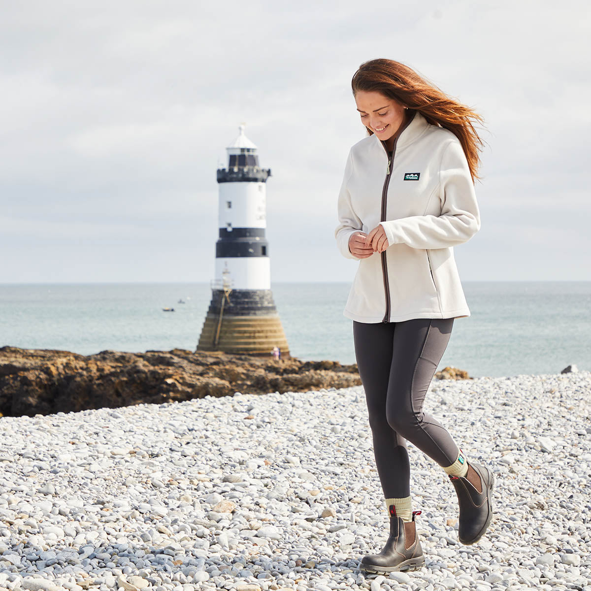 Woman walking on a pebbly beach with a lighthouse in the background. Wearing a Cream Windproof Fleece Jacket