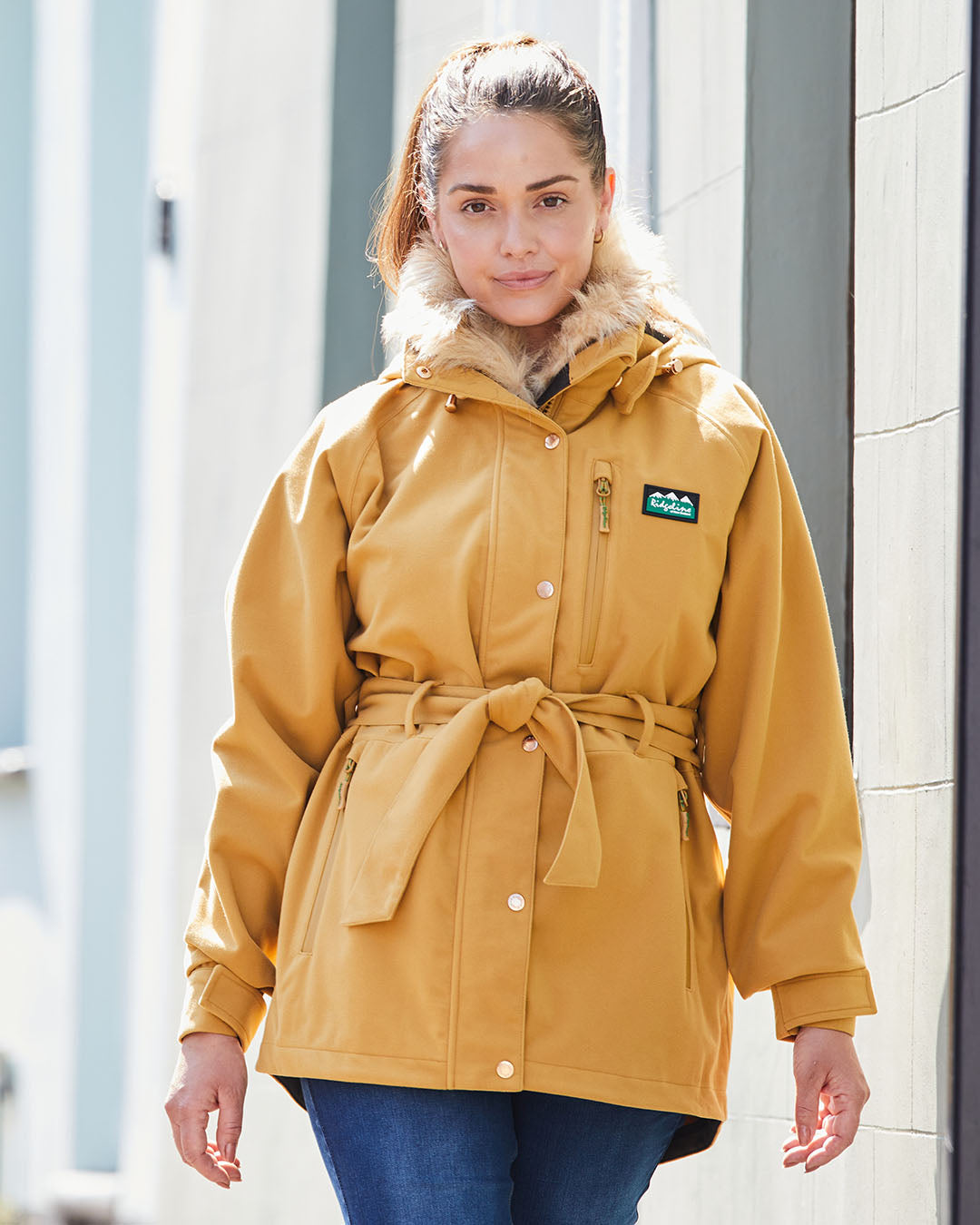 Woman wearing a mustard yellow coat with a brand logo, standing outdoors.