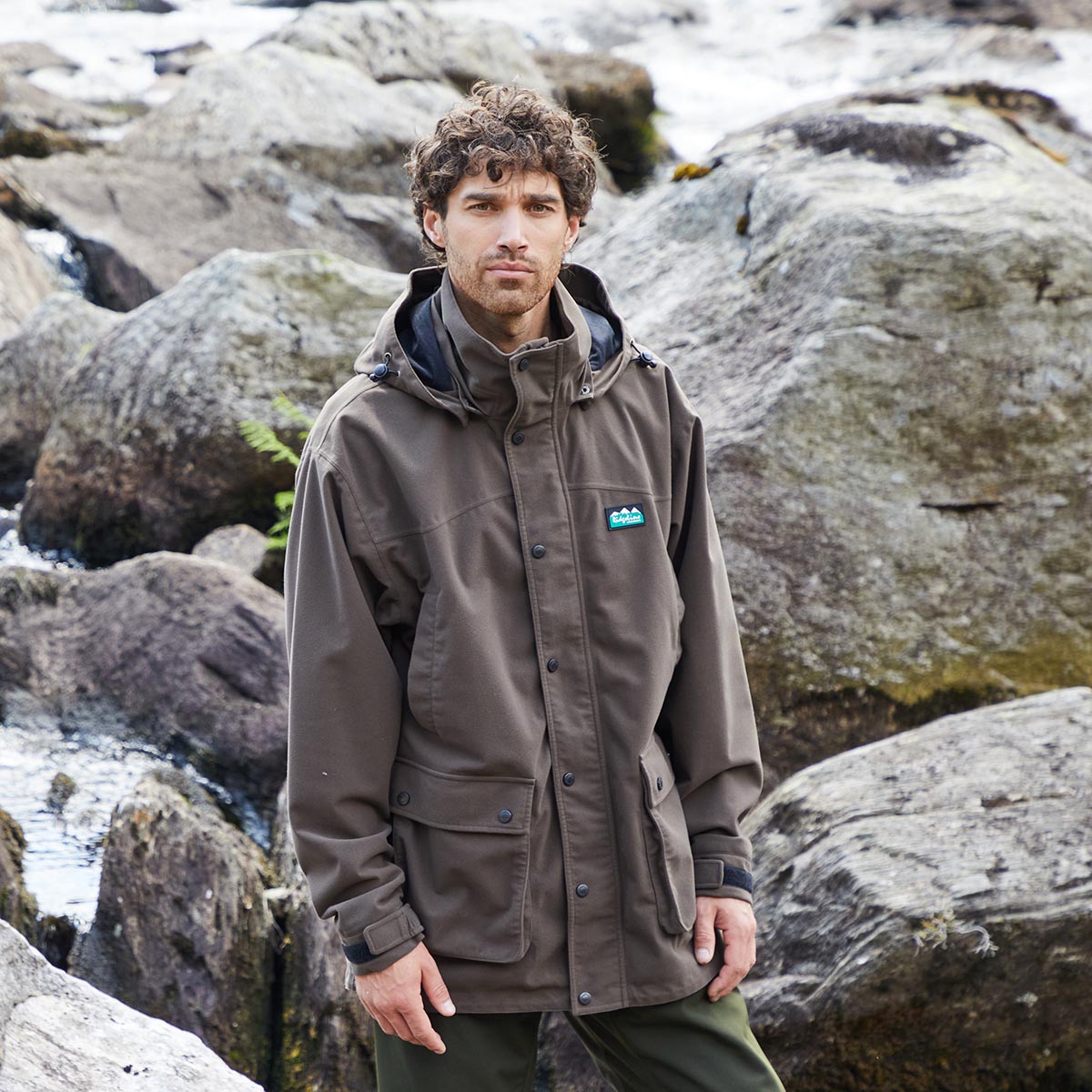 Man wearing a brown waterproof jacket with a Ridgeline Clothing logo standing on rocky terrain