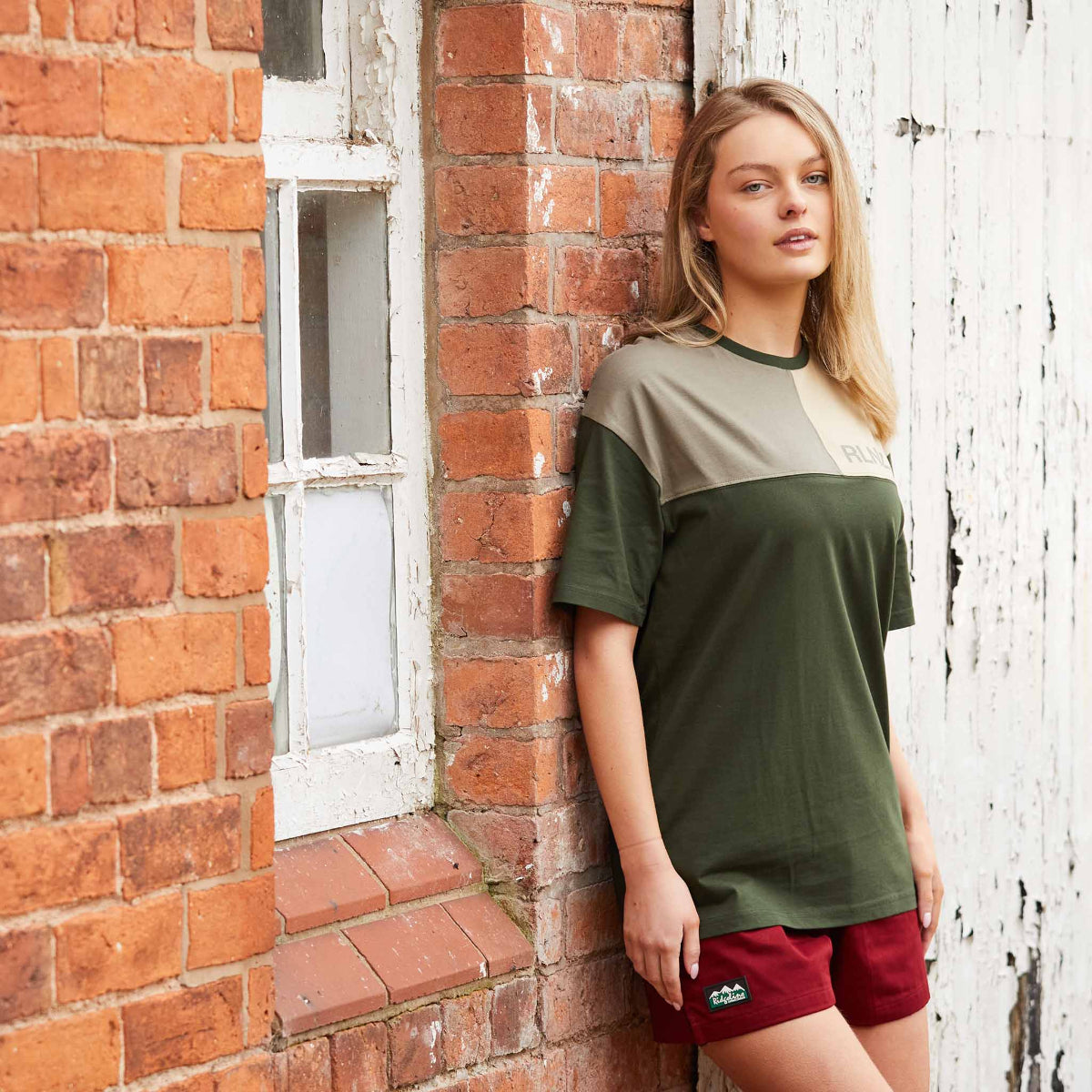 women wearing red shorts showing the Ridgeline badge with a olive green t-shirt the background shows a brick wall and a white fence. 