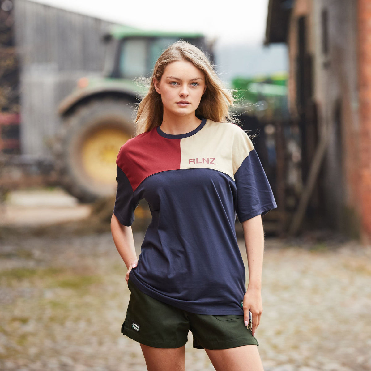 Woman wearing a colourful t-shirt with 'RLNZ' branding in an outdoor setting with a tractor in the background.