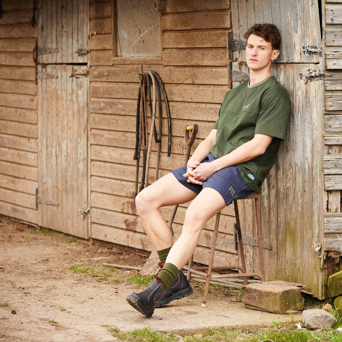 Man sitting on a stool against a wooden shed wearing green t-shirt and blue shorts.