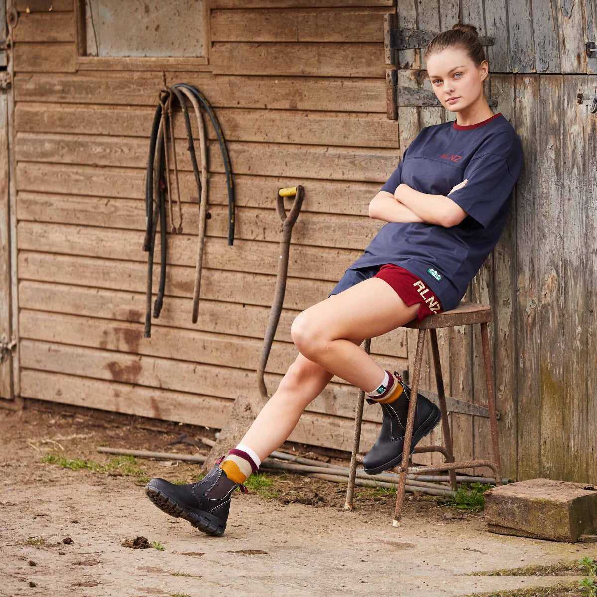 Woman sitting on a stool against a wooden wall with tools, wearing a navy shirt and red shorts.