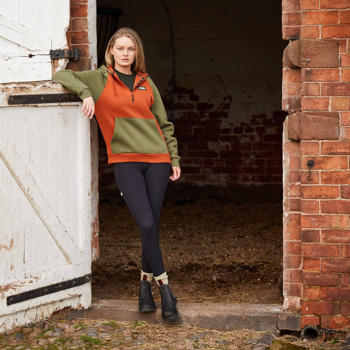 Woman wearing a pair of black leggings with a high waist and side pockets. Leaning against horse stable