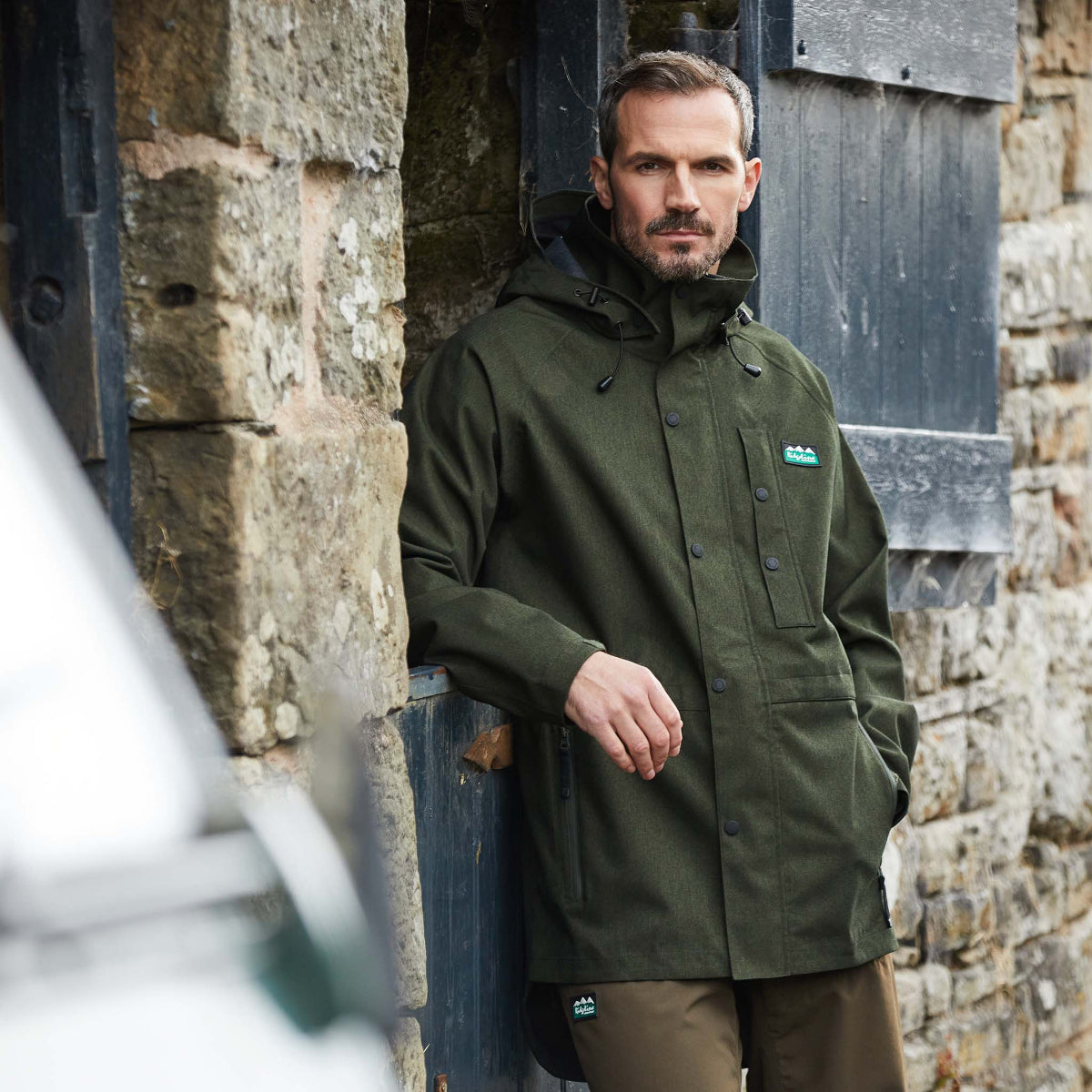 Man wearing a green Monsoon light waterproof jacket Windproof with a hood, featuring a full-length front zip, two zipped hand pockets, and a zipped chest pocket. standing & leaning outside a horse stable