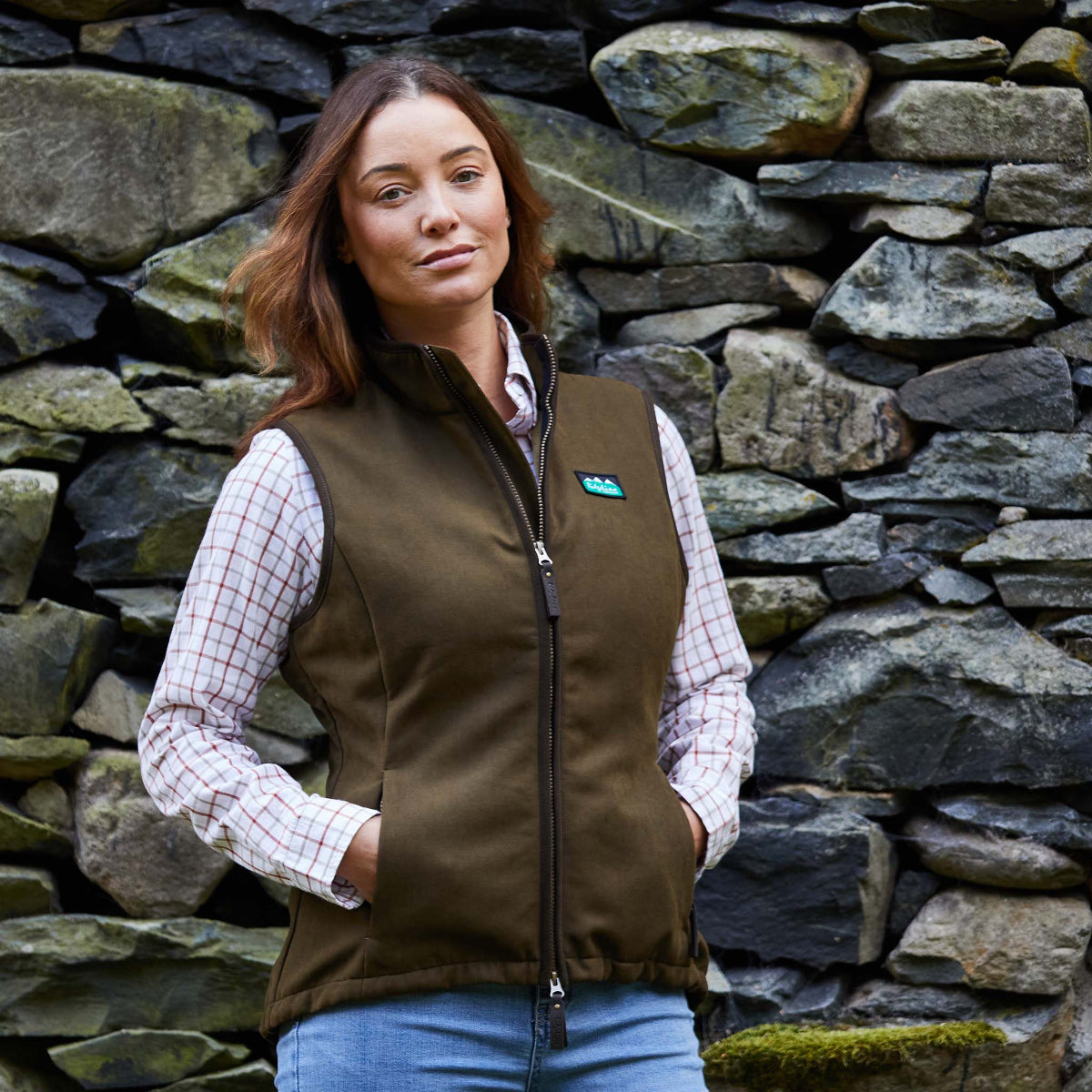 Woman wearing a brown vest gilet over a plaid shirt against a stone wall.