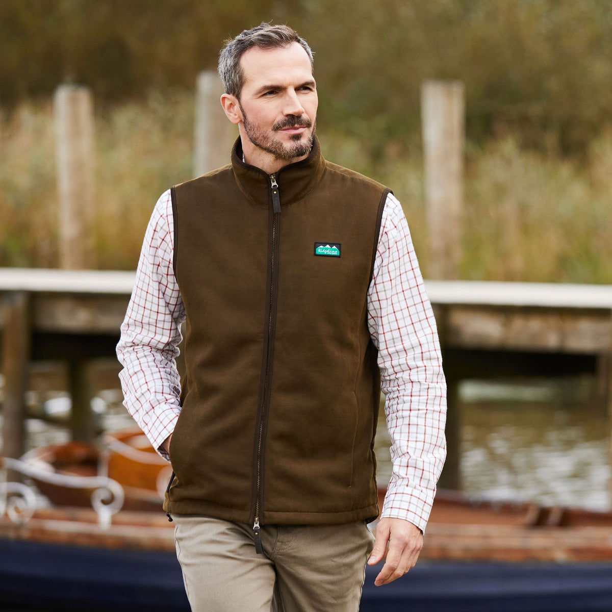 Man wearing a brown gilet vest over a plaid shirt with a blurred background of water and wooden posts.