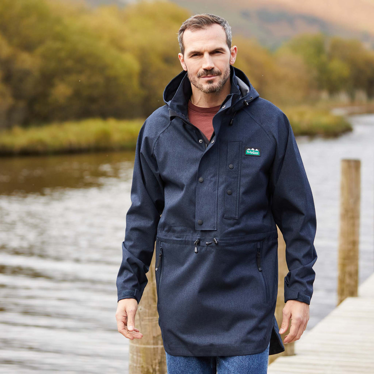 Man wearing a navy waterproof Smock standing on a wooden dock by a body of water with trees in the background.