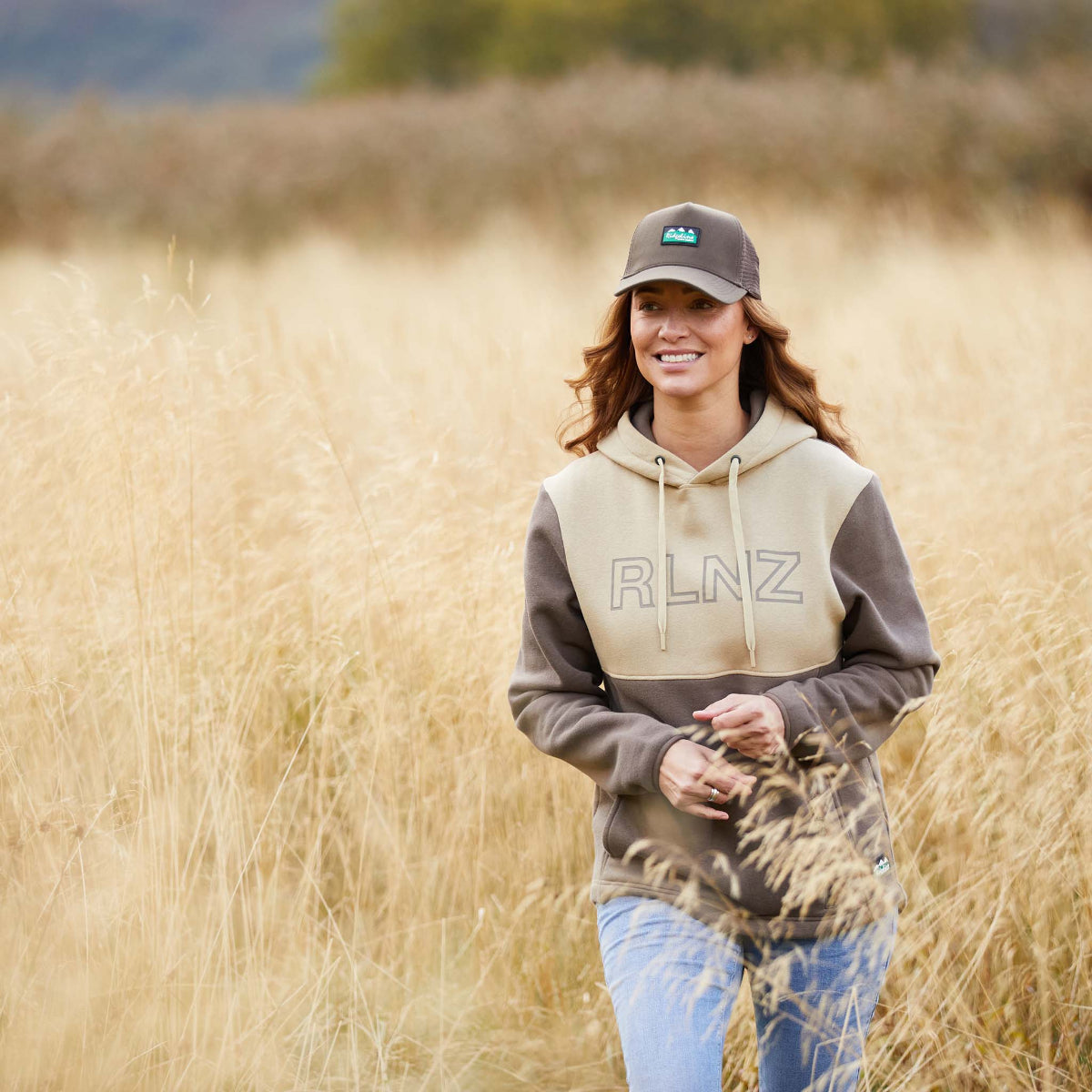 Woman wearing a two-tone brown and beige hoodie with a hood, kangaroo pocket, and the brand name 'RLNZ' printed across the chest. walking through field of wheat