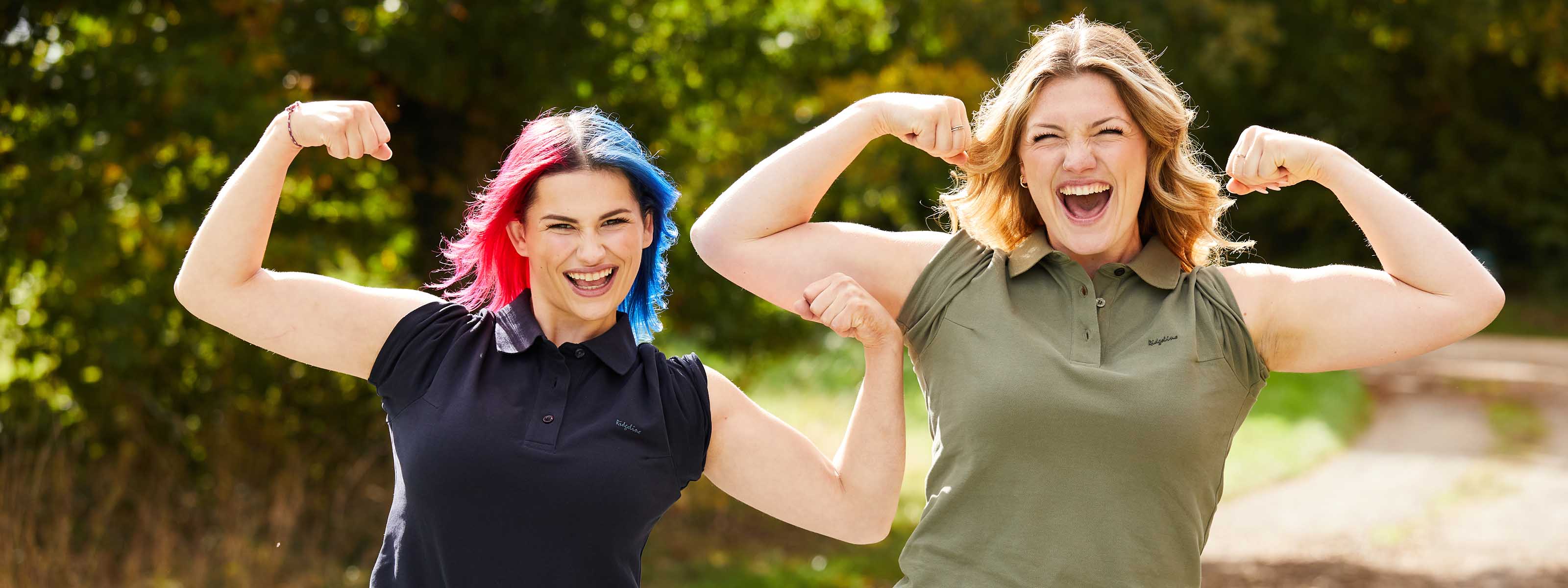 Two women flexing their muscles outdoors with a natural background