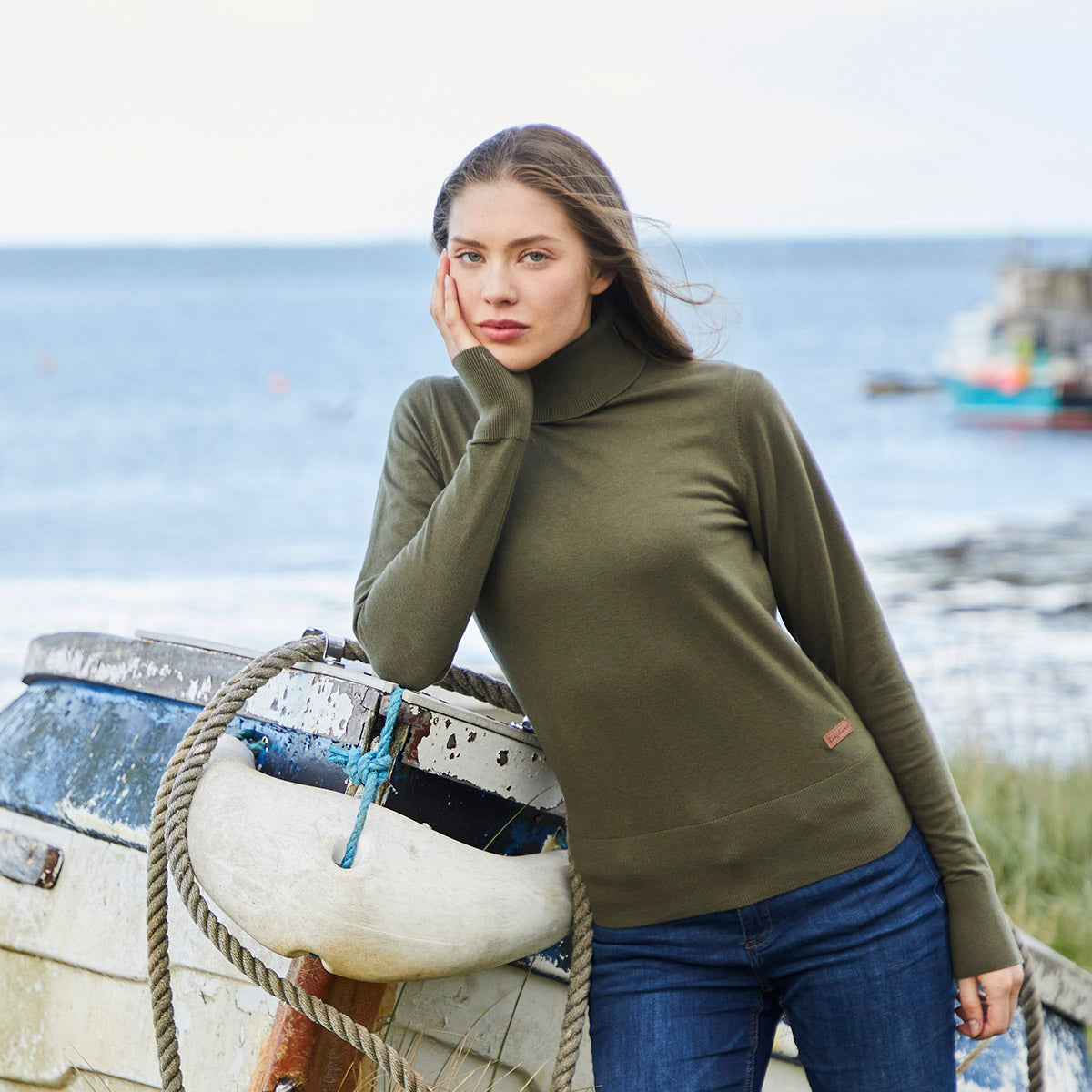 Lifestyle image of a model wearing a Ridgeline knitted roll neck pullover in olive green, featuring a fine-knit fabric, high roll-neck collar, and long sleeves. The jumper is styled with blue jeans and photographed beside a weathered boat on a coastal shoreline, showcasing its smart yet versatile outdoor look.

