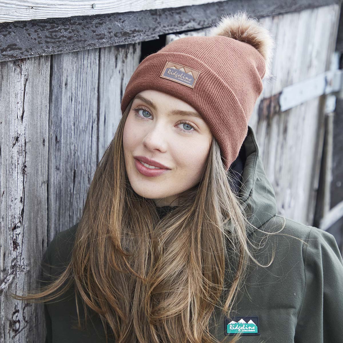 Lifestyle image of a person wearing a rust-brown ribbed knit beanie with a folded cuff and a large faux-fur pom-pom on the crown. A brown faux-leather Ridgeline of New Zealand logo patch is displayed on the front cuff. The person is dressed in a dark green outdoor jacket featuring a Ridgeline logo badge on the chest and is standing beside a weathered wooden fence. The outdoor setting and close-up composition highlight the beanie's soft knit texture, warm fit, and stylish cold-weather design.