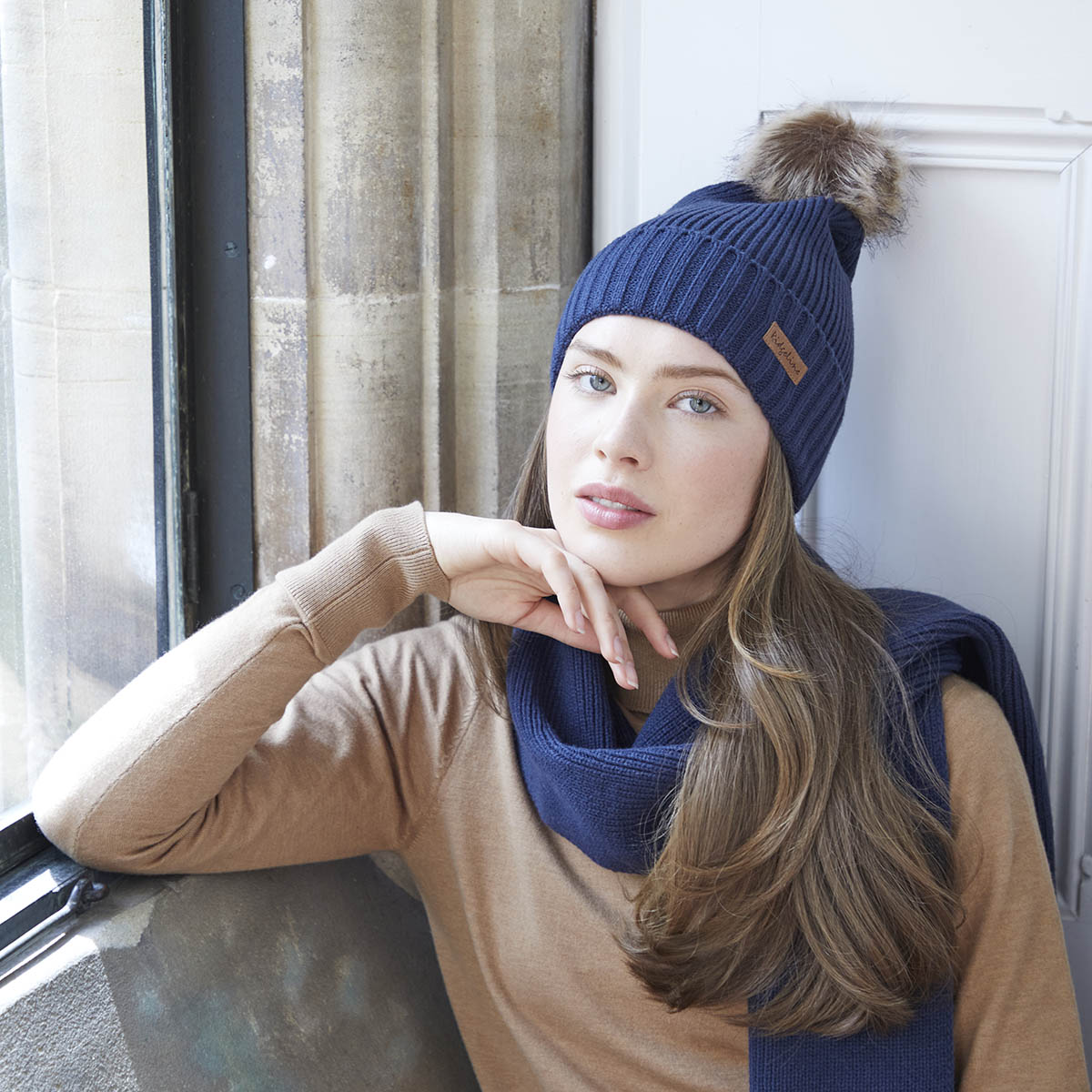 Model wearing navy ribbed knit beanie and scarf set with faux fur pom pom,  by window