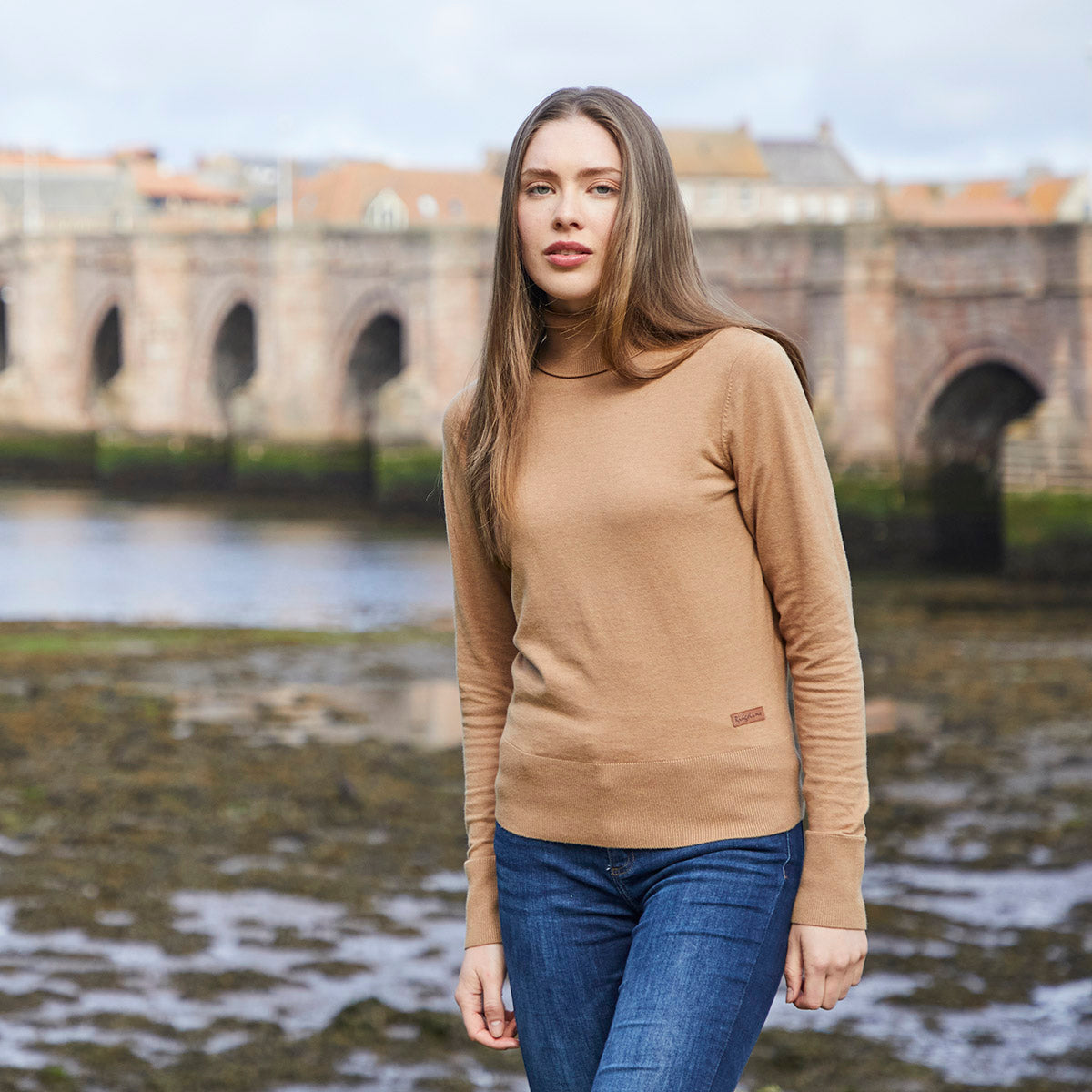Lifestyle image of a model wearing a Ridgeline knitted roll neck pullover in camel brown. The fine-knit jumper features a high roll-neck collar, long sleeves, and a ribbed hem, finished with a small Ridgeline logo tag near the waist. Styled with blue jeans and photographed outdoors by a riverside with a historic stone bridge in the background.
