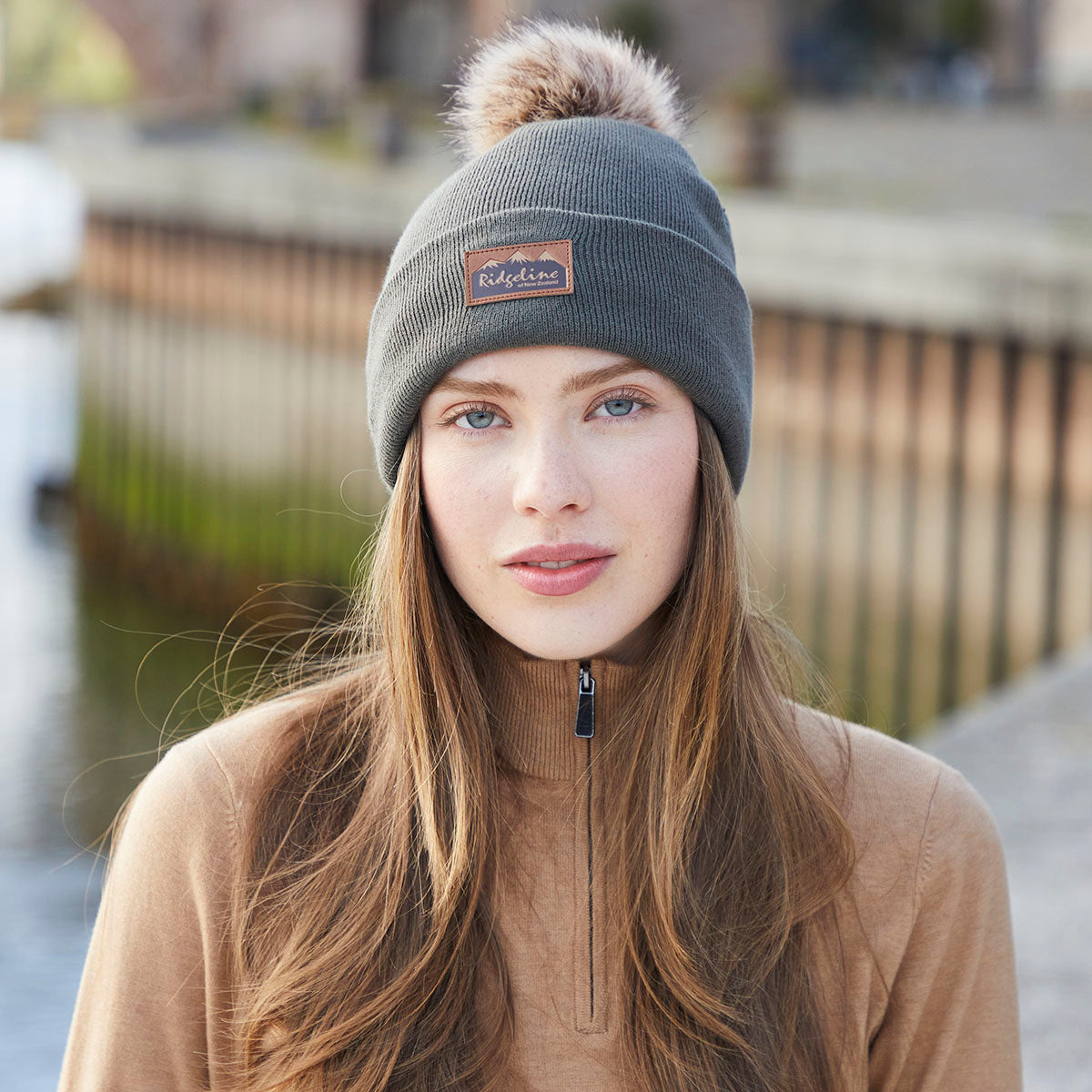 Lifestyle image of a person wearing a greenish-grey ribbed knit beanie with a folded cuff and a large faux-fur pom-pom on the crown. A brown faux-leather Ridgeline of New Zealand logo patch is displayed on the front cuff. The person is dressed in a light brown quarter-zip fleece top, and the image is taken outdoors with a softly blurred background featuring railings and natural tones. The photograph highlights the beanie's textured knit construction, warm fit, and stylish pom-pom detail for 