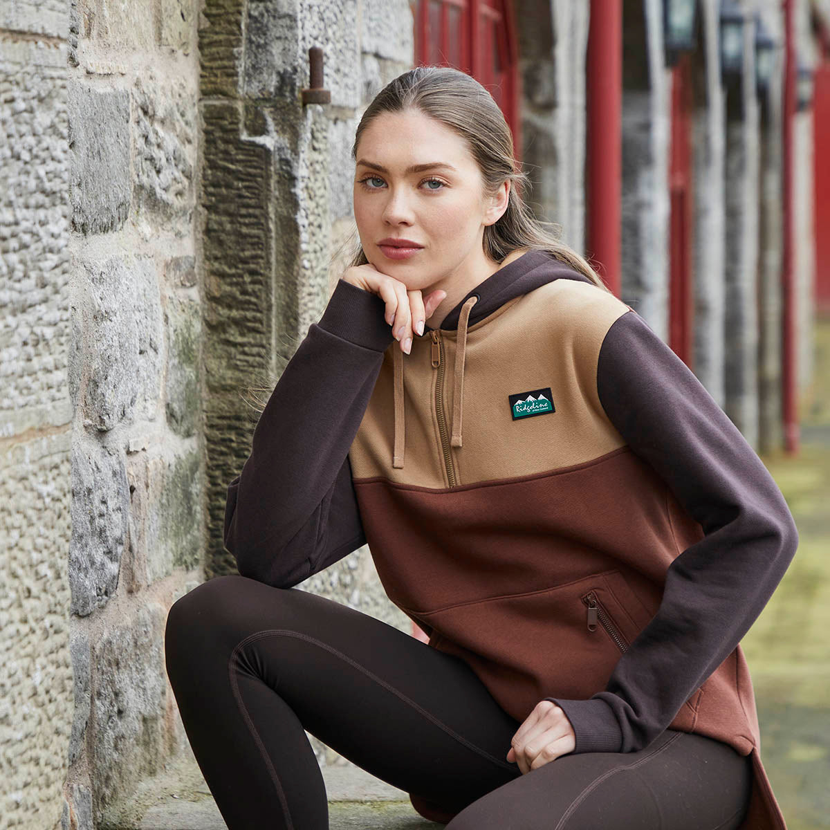 Lifestyle image of a person wearing a Ridgeline colour-block hoodie in beige, rust brown, and dark brown tones, paired with dark leggings. The hoodie features an adjustable drawstring hood, a full-length front zip, a Ridgeline logo patch on the chest, and a zipped front pocket for secure storage. The person is crouching beside a rustic stone wall with red-painted metal posts in the background, showcasing the hoodie in an outdoor setting.