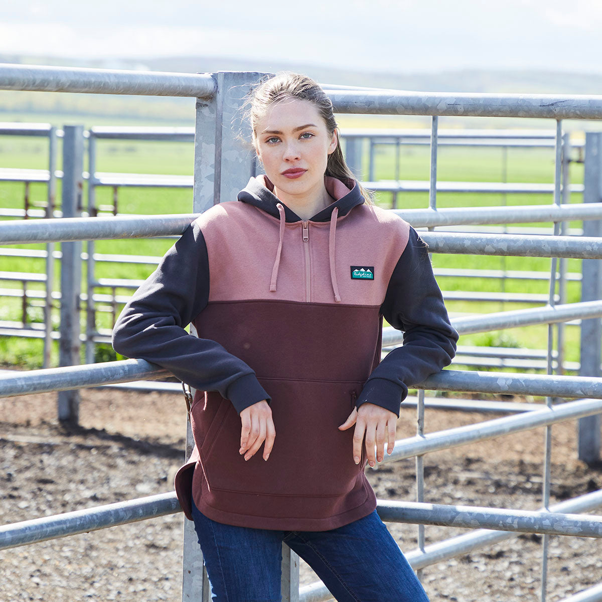 A person wearing a Ridgeline colour-block heavyweight hoodie in pink, brown and black with a 1/4 zip front and adjustable drawstring hood, standing beside metal livestock fencing outdoors and paired with blue jeans.
