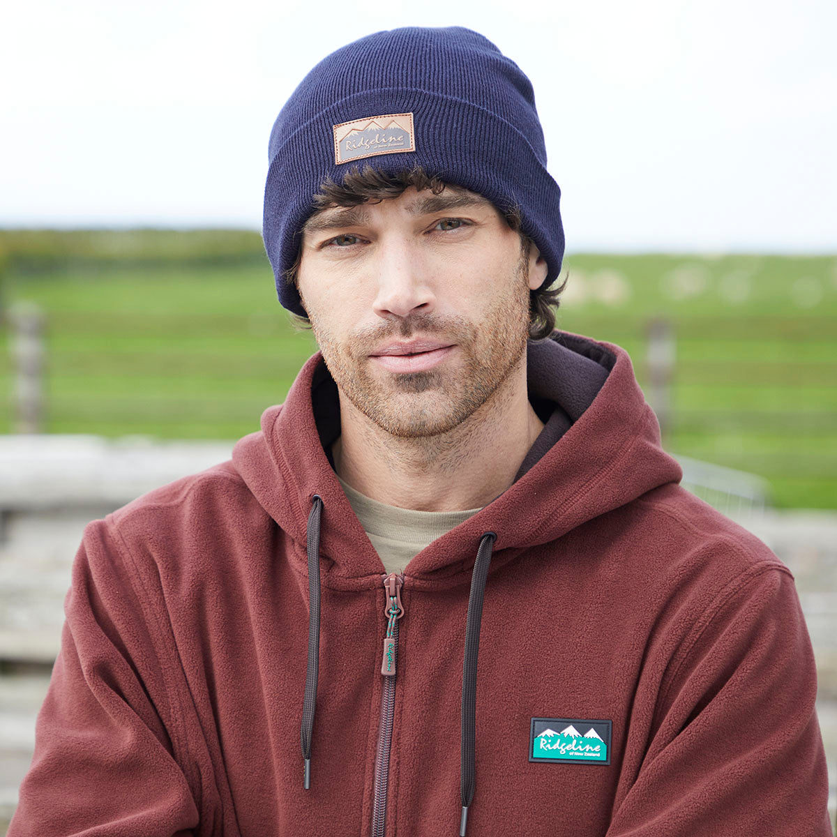 Lifestyle image of a person wearing a navy blue ribbed knit beanie with a folded cuff and a brown Ridgeline logo patch on the front. The person is also wearing a burgundy zip-up hoodie with a Ridgeline chest logo patch. The photo is taken outdoors with a softly blurred countryside background featuring green fields, showcasing the beanie in a casual, everyday setting.
