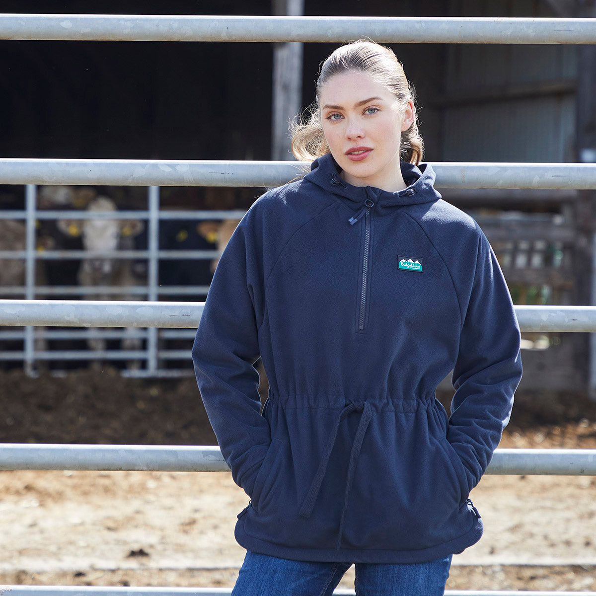 A woman wearing a navy blue pullover hoodie with a small green mountain logo on the left chest, standing in front of a metal fence.