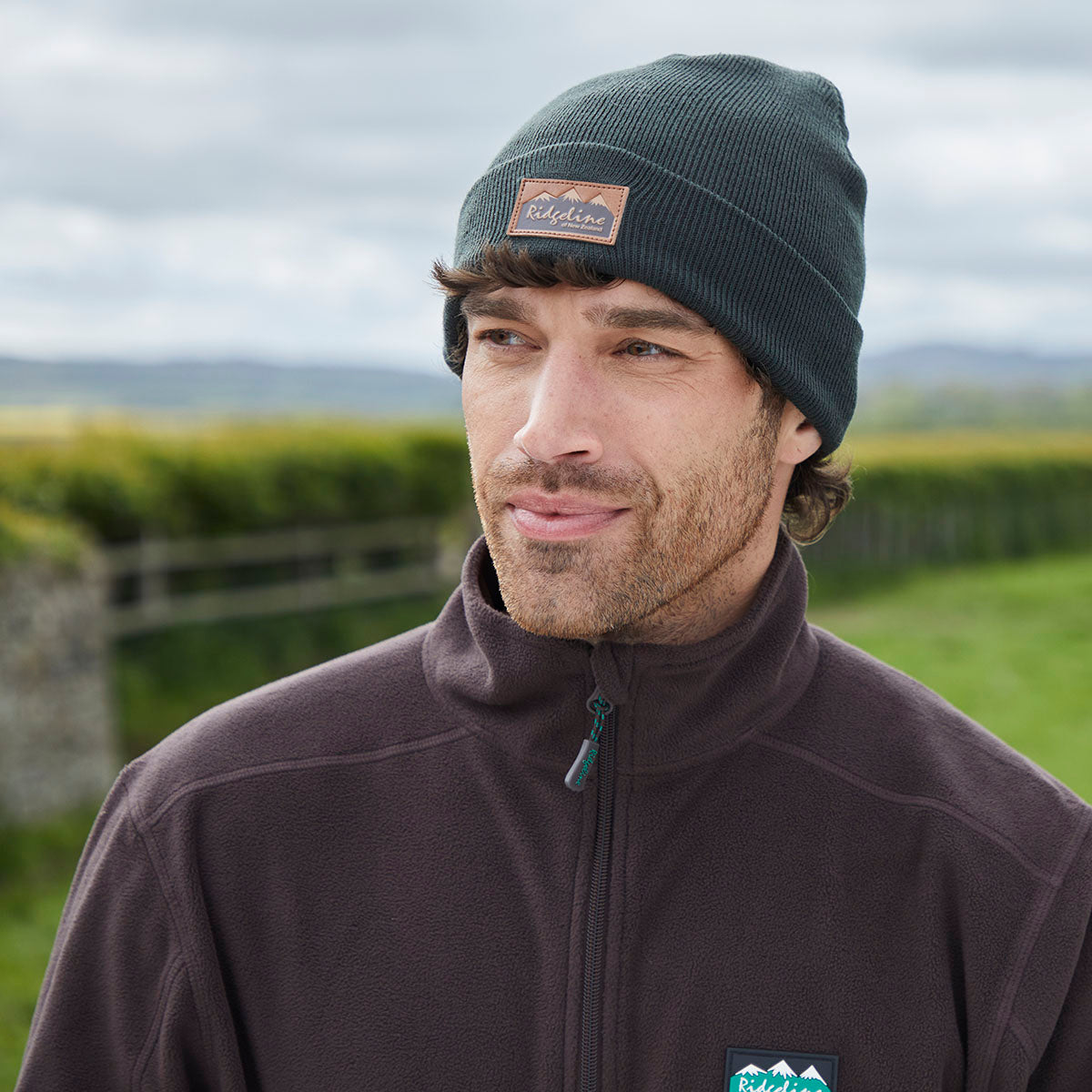 Lifestyle image of a person wearing a dark greenish-blue ribbed knit beanie with a folded cuff and a brown Ridgeline of New Zealand logo patch on the front. The person is dressed in a dark zip-up fleece jacket featuring a Ridgeline logo badge on the chest. The photograph is taken outdoors with a softly blurred countryside backdrop of green fields, hedgerows, and an overcast sky, showcasing the beanie's comfortable fit, knit texture, and practical outdoor style.
