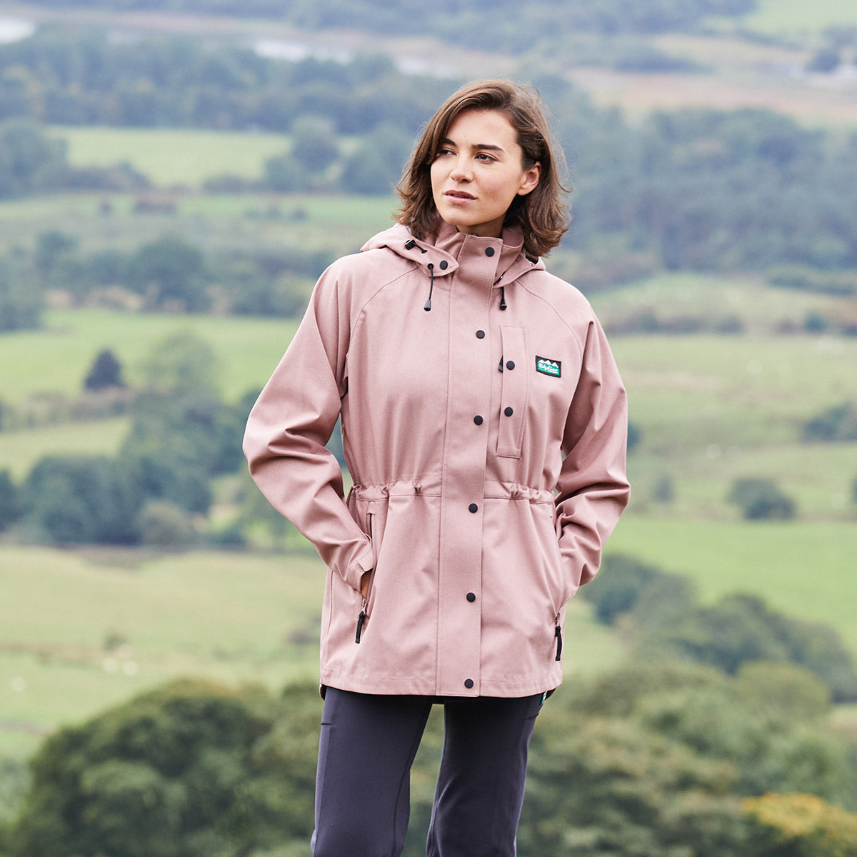 Person wearing a pink raincoat standing in a scenic outdoor setting with green fields and trees.