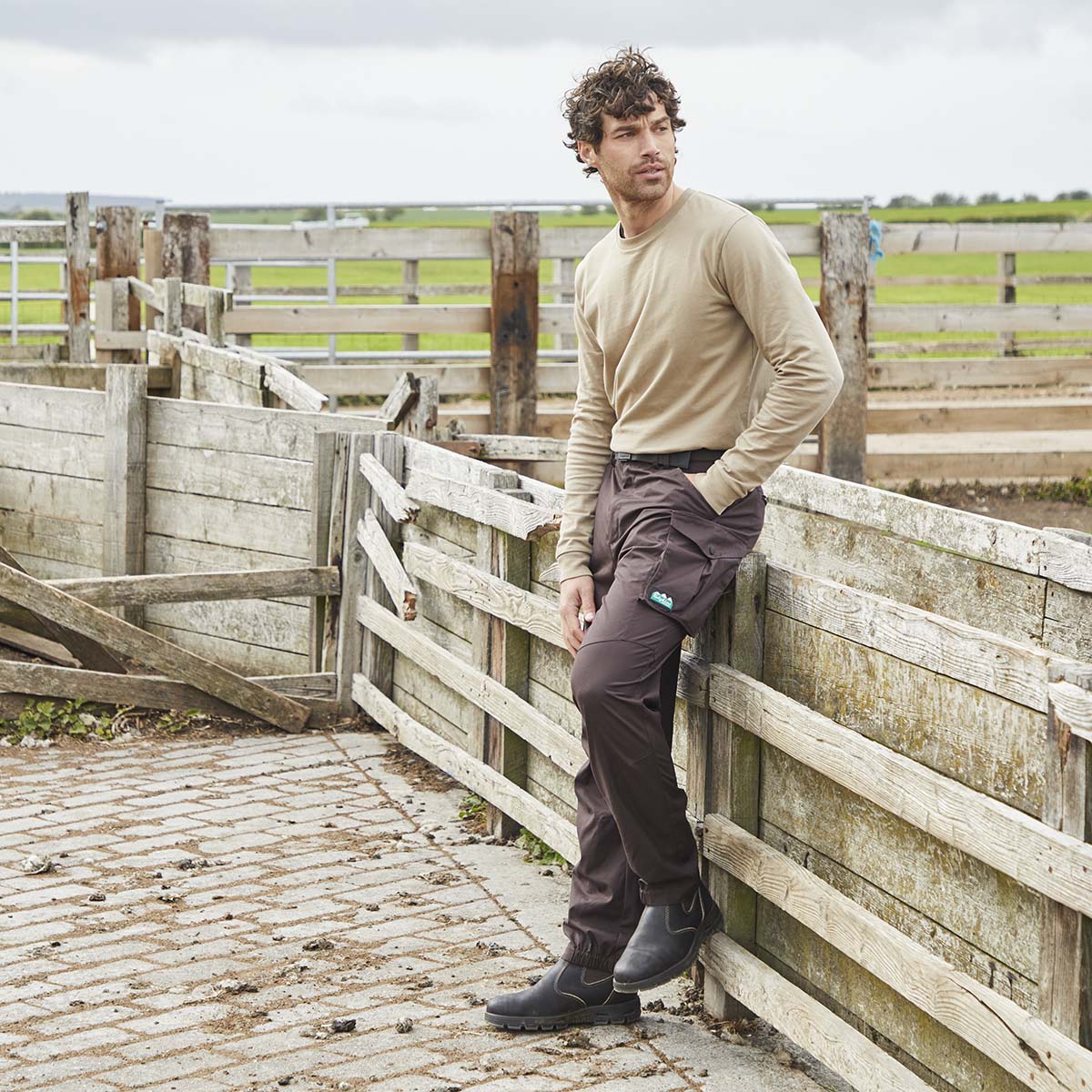 Beige long sleeve top with brown cargo trousers and dark ankle boots, worn outdoor model view by wooden fence in rural setting
