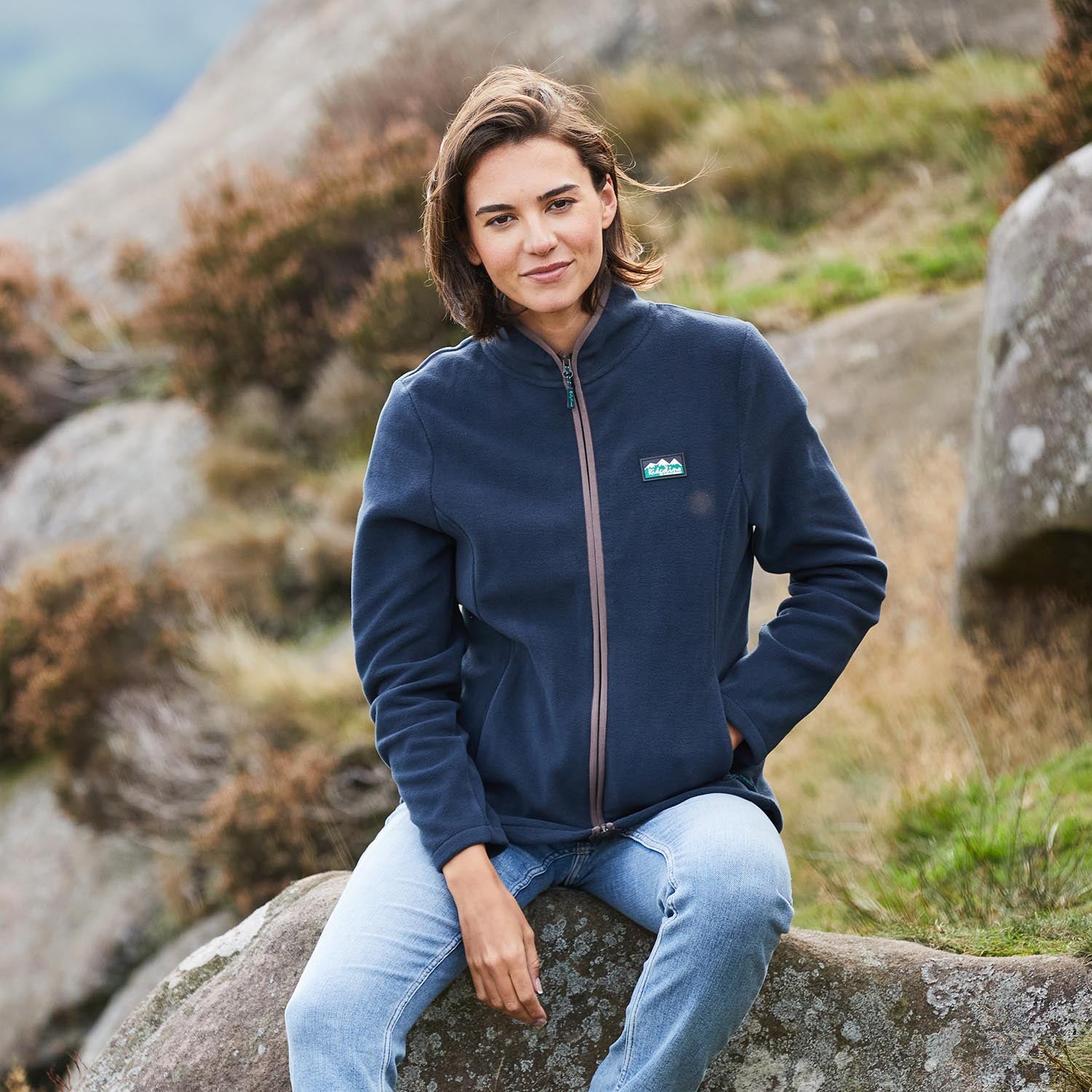 Woman wearing a blue jacket with a logo, sitting on a rock in a natural setting.