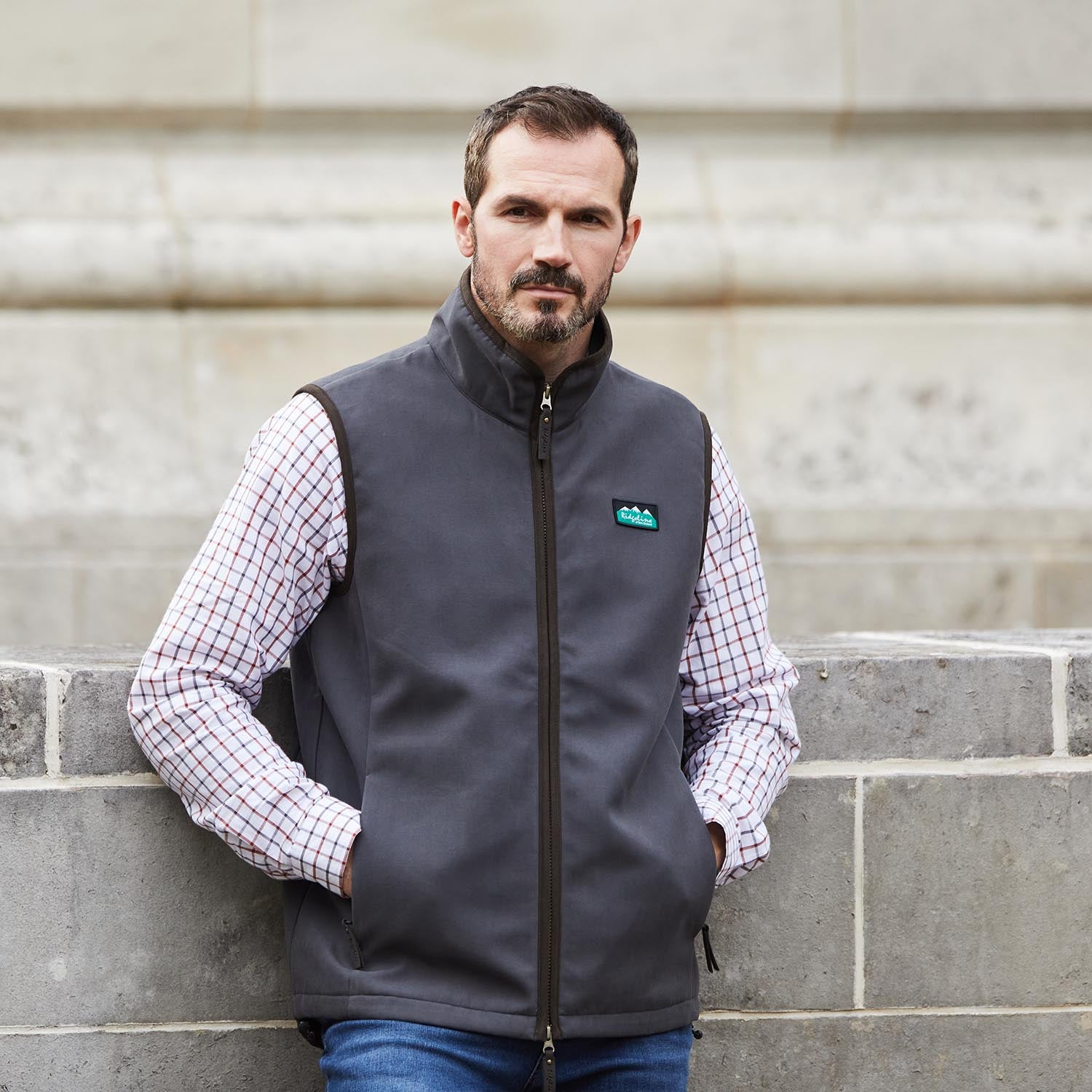 Man wearing a gray vest with a logo, standing against a stone wall.