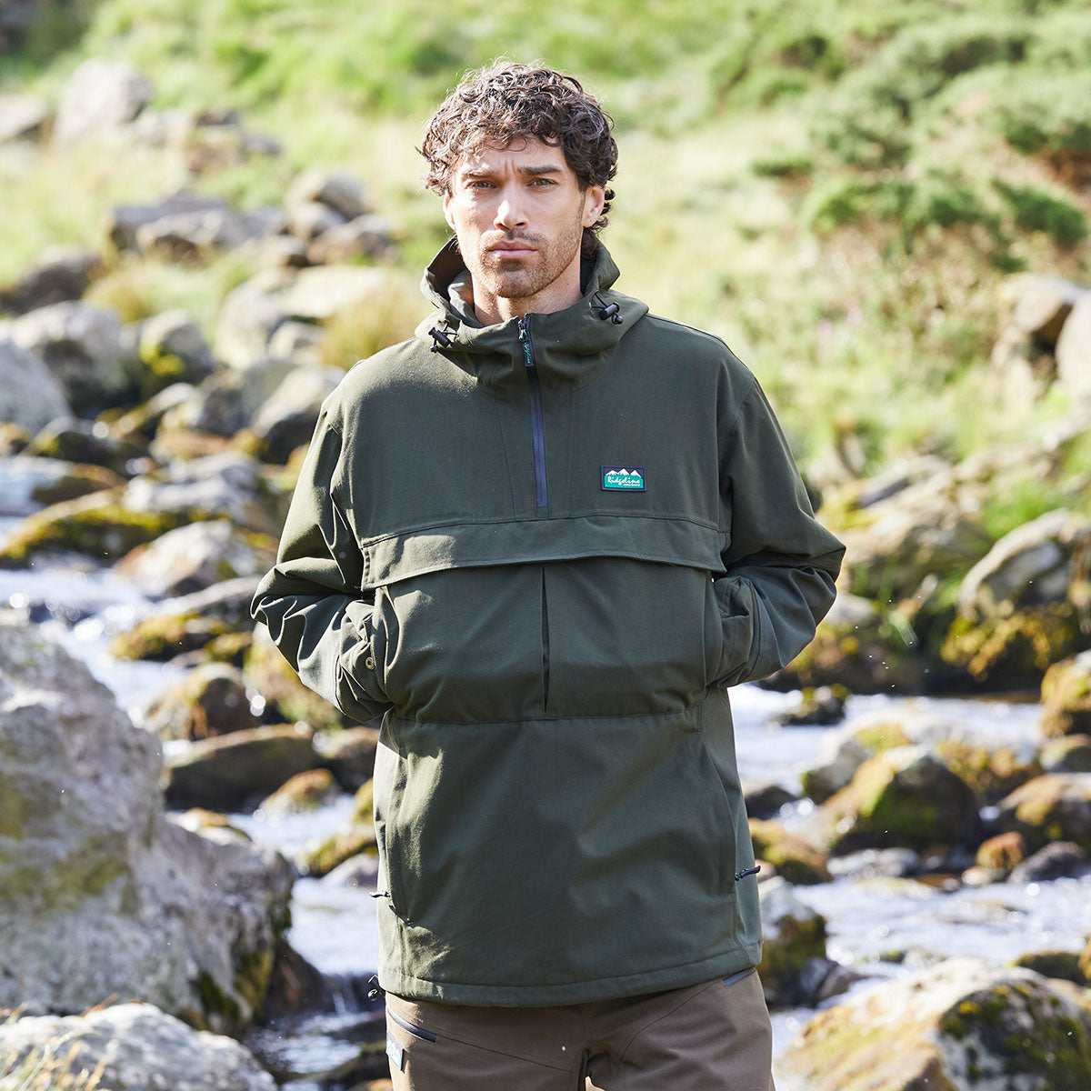 Olive green hooded overhead jacket with half zip front and chest pocket, outdoor model view by stream